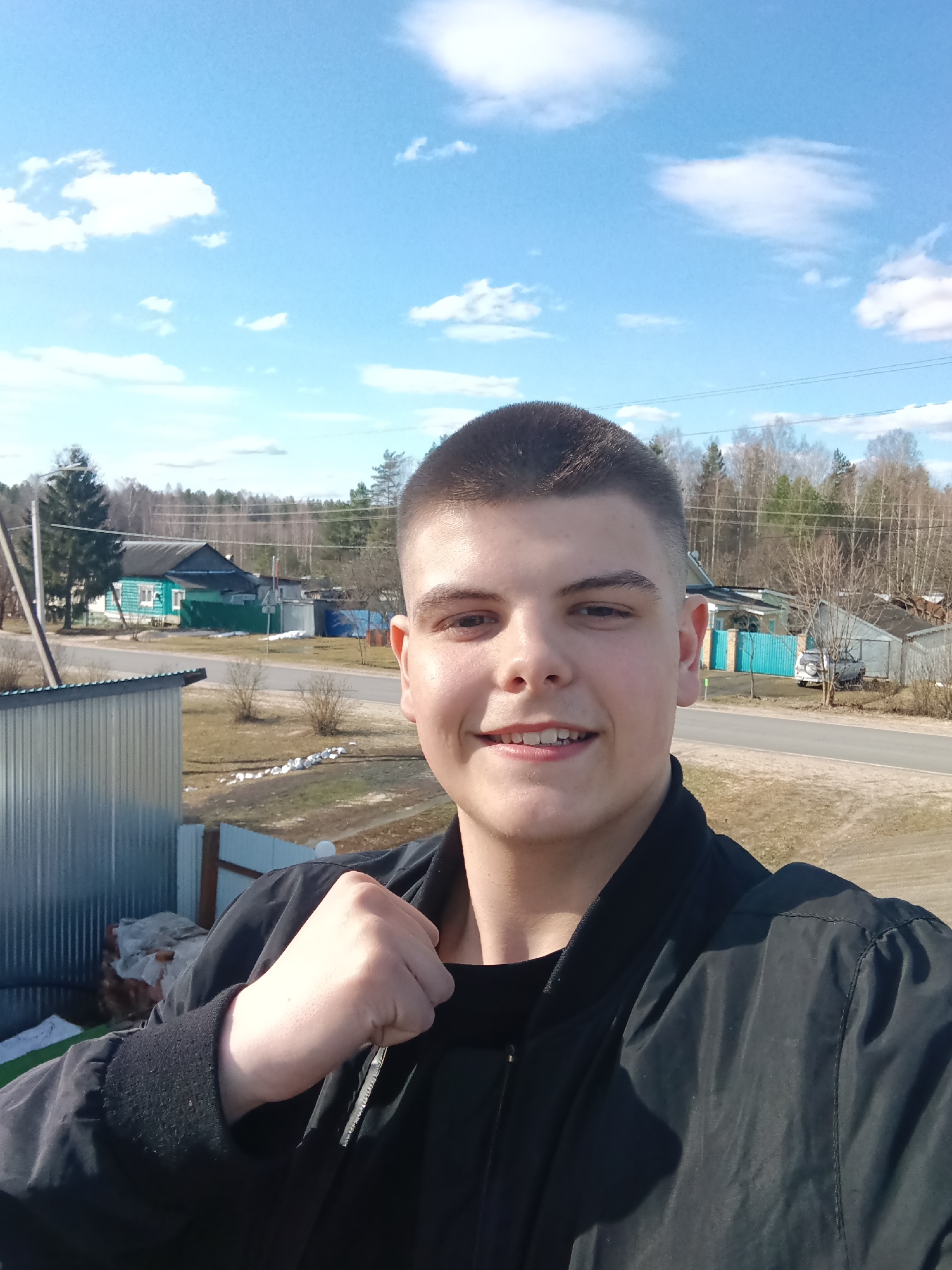 A young man is taking a selfie outdoors on a sunny day in Красный Маяк, Russia. The man has short, dark hair, fair skin, and is wearing a black jacket over a black shirt. He is smiling slightly. Behind him is a view of a rural or suburban scene in Красный Маяк, Russia, including houses, trees, a road, and a blue and cloudy sky. The ground is mostly brown, suggesting it is early spring. There is a corrugated metal structure on the left side of the frame, possibly a fence or building.