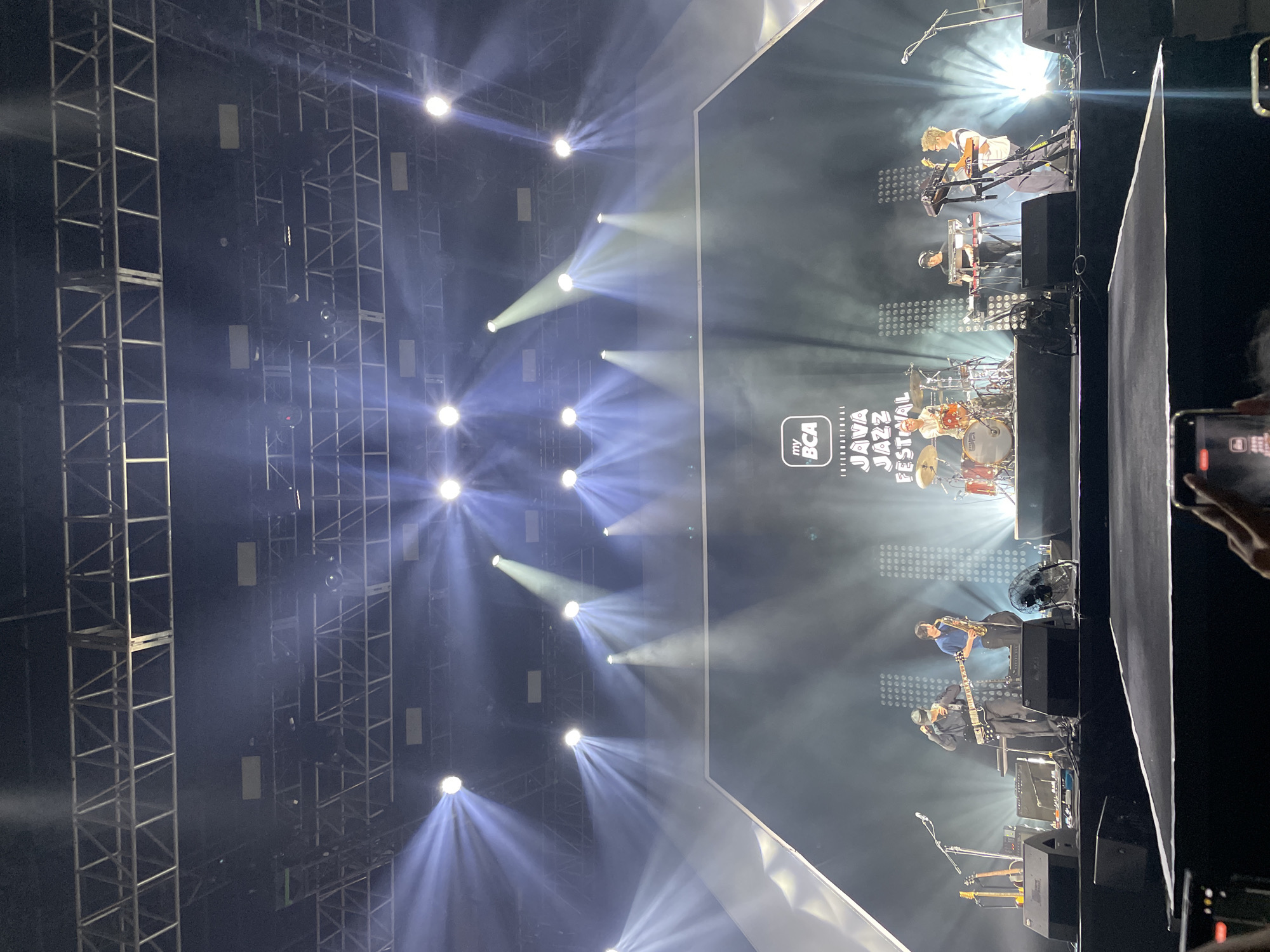 The image depicts a live music performance at the 2023 Java Jazz Festival, located in Salembaran, Indonesia. A band is on stage, lit by numerous spotlights, in front of a large screen displaying the festival logo. The band includes a drummer, keyboard players, a saxophonist, and a bassist. A metal truss structure supporting the lighting is visible. A person can be seen in the foreground, holding a mobile phone, likely recording the performance.