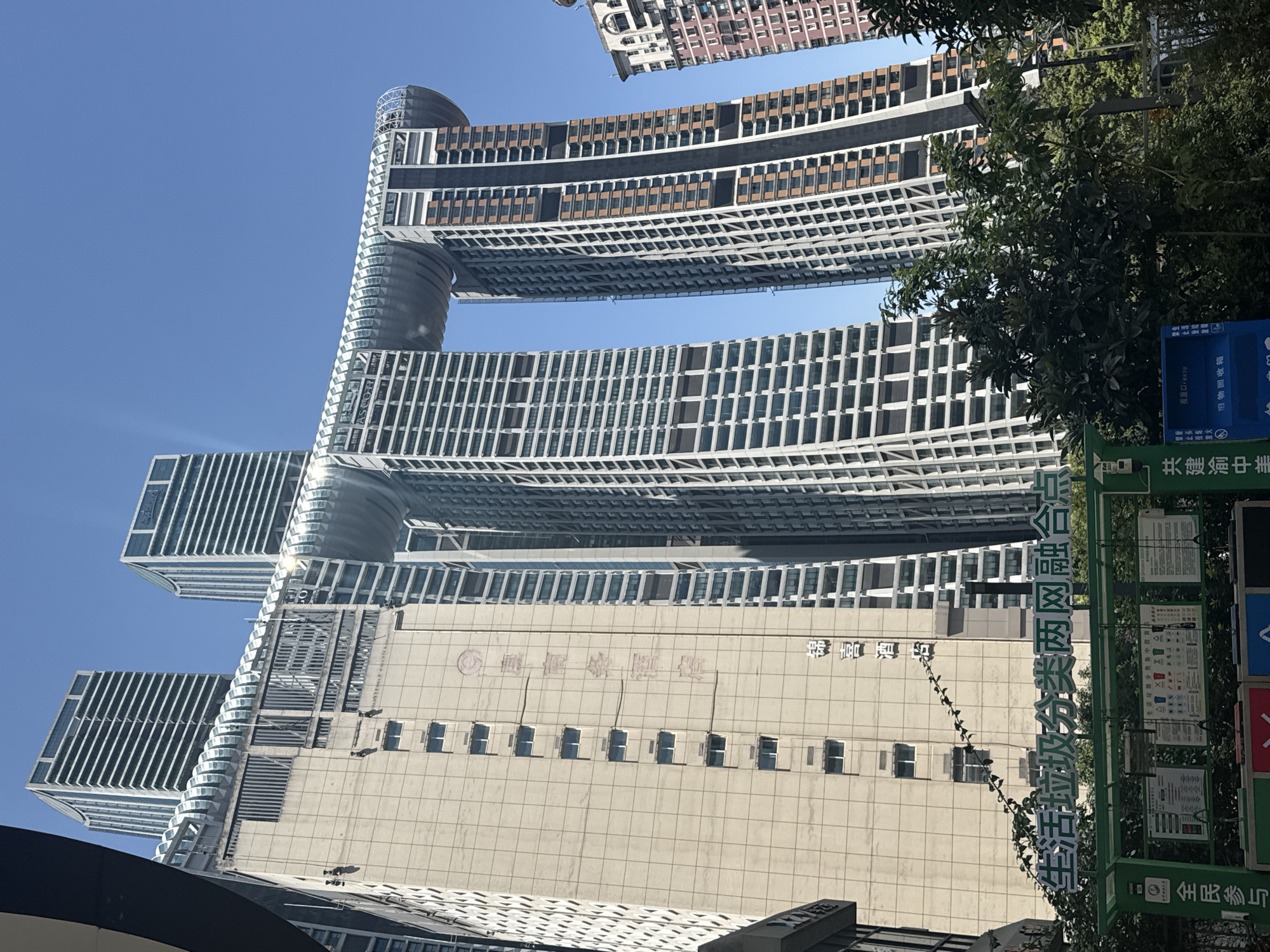 The image shows a street view of the Chongqing Raffles City in Yuzhong District, China. The iconic building, designed by architect Moshe Safdie, features a horizontal skybridge connecting four towers, resembling a ship or a dragon. The skybridge has a transparent, curved facade and is illuminated by the sun. Two of the towers are significantly taller and narrower, with a distinctive diagonal grid pattern on their facades. Another wider, older-looking building is partially visible to the left, with rows of small windows and vertical Chinese characters printed on its beige facade. In the foreground, a green structure with signs related to waste sorting and recycling is present, with trees partially obscuring the view. The sky is clear and blue. The overall scene is an urban environment dominated by modern architecture.