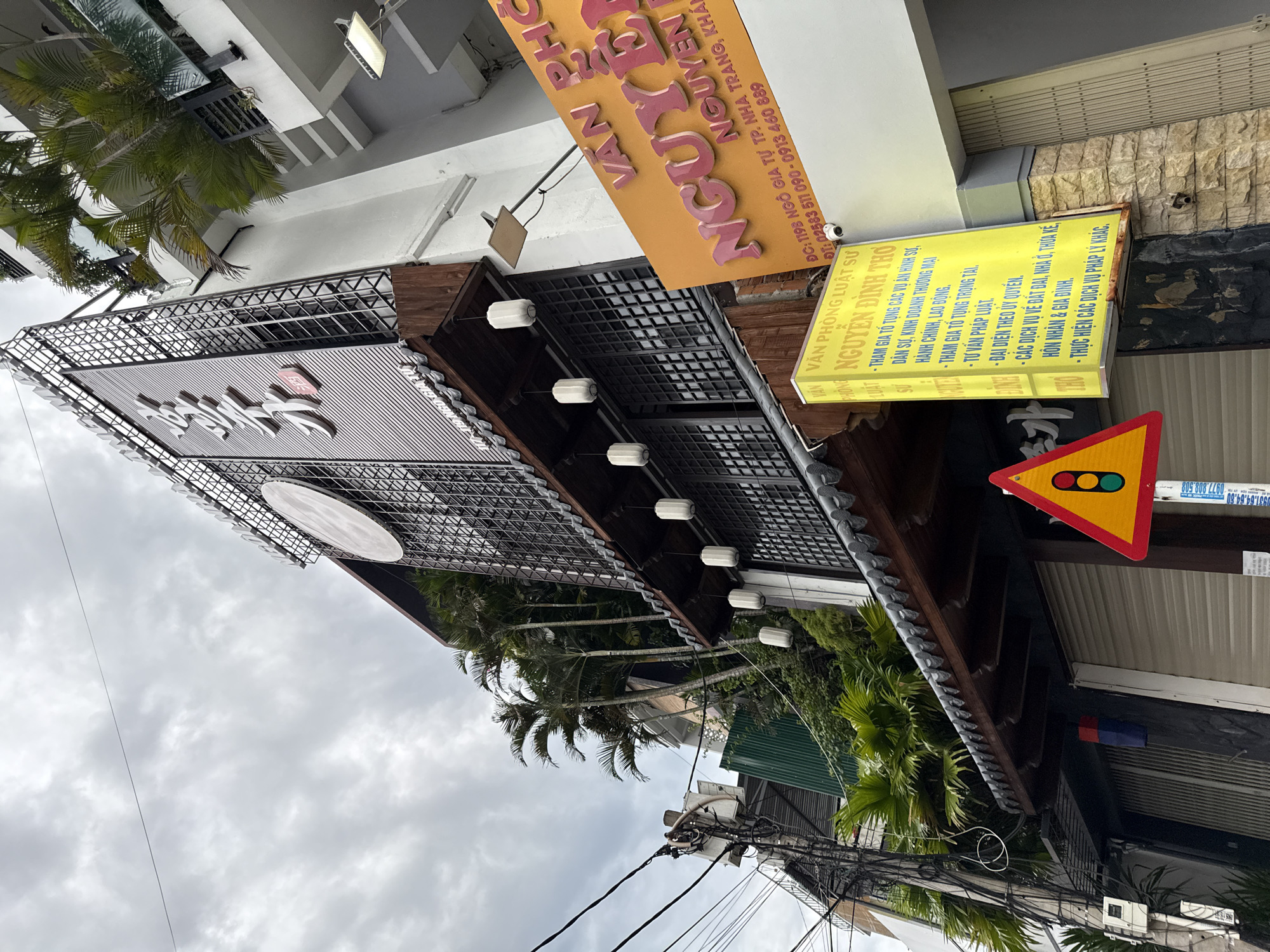 The image depicts a street scene in Nha Trang, Vietnam, featuring a building with a distinctive architectural style. The building has several floors, with a prominent dark-colored, tiled roofline reminiscent of traditional East Asian design. The facade is adorned with metal lattice work and what appears to be vertical wooden slats. Large, white, cylindrical lanterns hang from the eaves. On the building's upper section, large white characters are mounted, possibly indicating the name of a business or establishment. Green foliage, including palm trees, is visible growing on balconies and terraces integrated into the building's structure.
To the right of the main building, a bright orange sign with large pink text reads "VAN PHO NGUYEN". Below this, smaller text in Vietnamese indicates an address: "DC: 119B NGO GIA TU TP. NHA TRANG, KHANH HOA". Further details on the sign include telephone numbers. Another yellow sign to the left also features Vietnamese text, listing legal services. A traffic light sign with red, yellow, and green lights is also visible in the foreground, positioned on a pole. The street itself is paved, and the overall impression is that of a bustling urban environment. The sky is overcast. There are no visible people in the image. The scene suggests a commercial area in the city of Nha Trang.