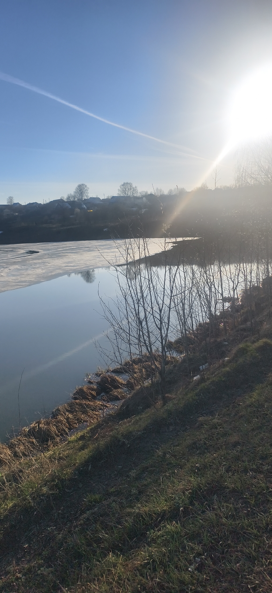 This landscape photograph presents a serene view of a partially frozen body of water in Фоки, Russia, bathed in the bright light of a low-hanging sun. The scene likely depicts late winter or early spring, given the ice on the water and the bare branches of the trees.
The sun, positioned on the upper right, creates a strong glare and lens flare, suggesting either early morning or late afternoon. The sky is clear and blue, marked by a distinct white contrail stretching diagonally across the upper left.
A significant portion of the water on the left side is covered with a sheet of ice, reflecting the clear sky. Towards the right, the water is open and calm, offering reflections of the surrounding landscape. The foreground features a gently sloping bank covered with sparse, dry grass and some emerging green growth. Bare, deciduous trees and bushes line the water's edge and the bank, their dormant branches reaching towards the sky.
In the background, across the expanse of water, several houses with dark roofs are visible on a hillside, indicating a rural village or settlement. The overall impression is one of quietude and the crisp atmosphere of a sunny, cold day. There are no people visible, and no discernible text is present in the image.