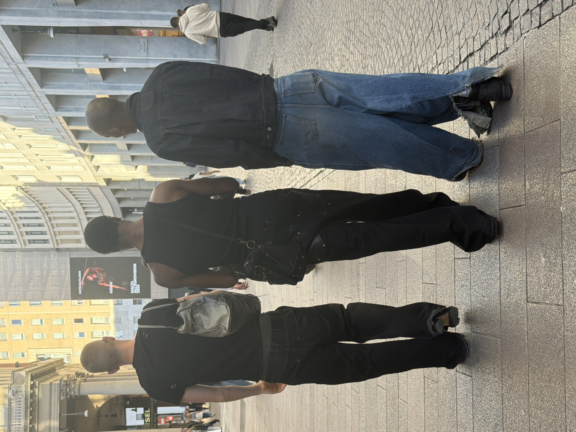 The image shows three people walking on a paved street in Milan, Italy. The first person is wearing a dark denim jacket and loose-fitting blue jeans with tattered edges. The second person is wearing a black tank top and black pants, with a black bag across their chest. The third person is wearing a black t-shirt, black pants, and carrying a gray bag over their shoulder. In the background, there is a billboard, buildings, and other pedestrians.