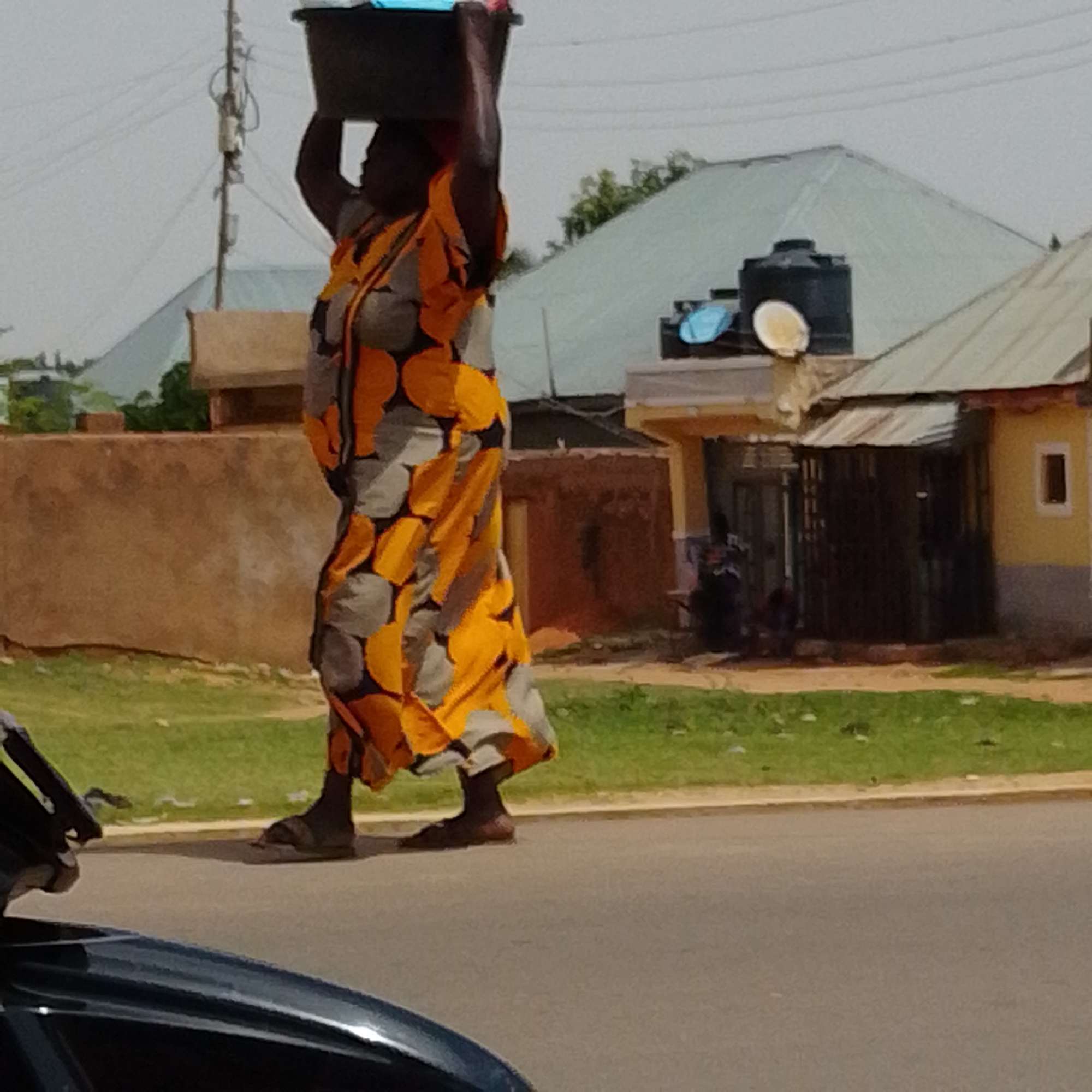 In Gombe, Nigeria, a person walks down a street carrying a large black basin on their head. The individual is wearing a vibrant, patterned dress with large orange and gray circles against a dark background. They are also wearing dark sandals. Behind the person, the environment includes a brown wall, a grassy area, a road, and several buildings. One building has a light green roof, and another is painted yellow with a dark metal gate. A black water tank is visible on top of one of the buildings. The sky is overcast. In the foreground, a portion of a dark vehicle is visible.