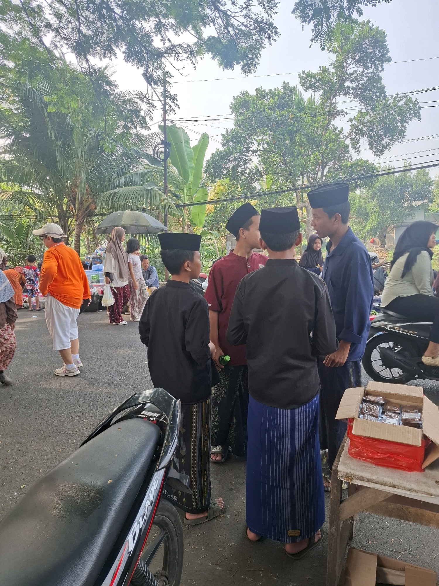 The image depicts an outdoor scene in Bojong, Indonesia, likely a street or public area, with several people present. In the foreground, a motorcycle is partially visible. A group of young men wearing black peci hats and sarongs are the central focus, with some appearing to be in conversation. One man in a dark blue shirt and sarong is looking towards another. To the left, a man in an orange shirt and white shorts stands with his back to the camera. In the background, other individuals and various objects are scattered, including what appears to be a market stall with an umbrella, a box of packaged goods, and another motorcycle. The environment is characterized by trees, overhead wires, and a paved surface, suggesting a semi-urban or residential setting. The lighting indicates it is daytime.