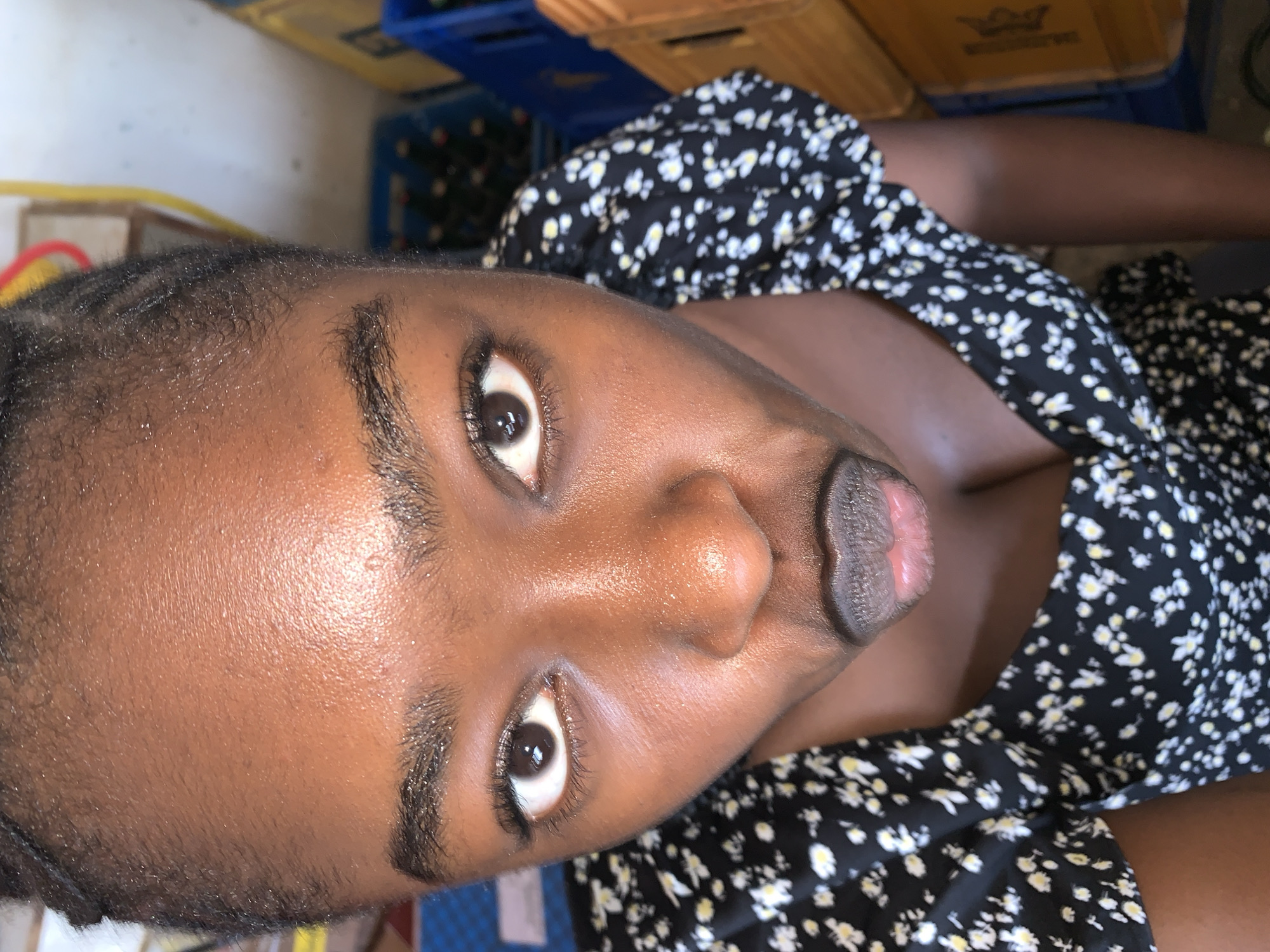 A close-up, rotated portrait features a person with dark skin, appearing to be a young female, looking directly into the camera. Her head and upper torso dominate the frame, with her body angled slightly away. Her skin has a visible sheen on the forehead and cheeks, indicating natural oils or sweat, with discernible pores. She has prominent, dark eyes with white sclera and visible eyelashes. Her eyebrows are dark and well-defined. Her lips are full, with a darker outer line and a lighter, pinkish inner part, and are slightly pursed. Her hair appears to be in short, possibly braided, styles around her hairline.
She is wearing a black dress patterned with small, scattered white and yellow daisy-like flowers. The garment seems casual.
The background, partially out of focus, reveals industrial-style storage crates. A blue plastic crate is visible to the left, and a brown crate, possibly wood or cardboard, is seen to the right, behind the subject's shoulder. On the brown crate, a faint golden emblem or logo with indistinct text or symbols is visible. These elements suggest an informal indoor setting, possibly a storage area or backroom, consistent with the New Stadium location in Nigeria. The lighting appears natural, suggesting it is daytime. The subject's expression is direct and composed.