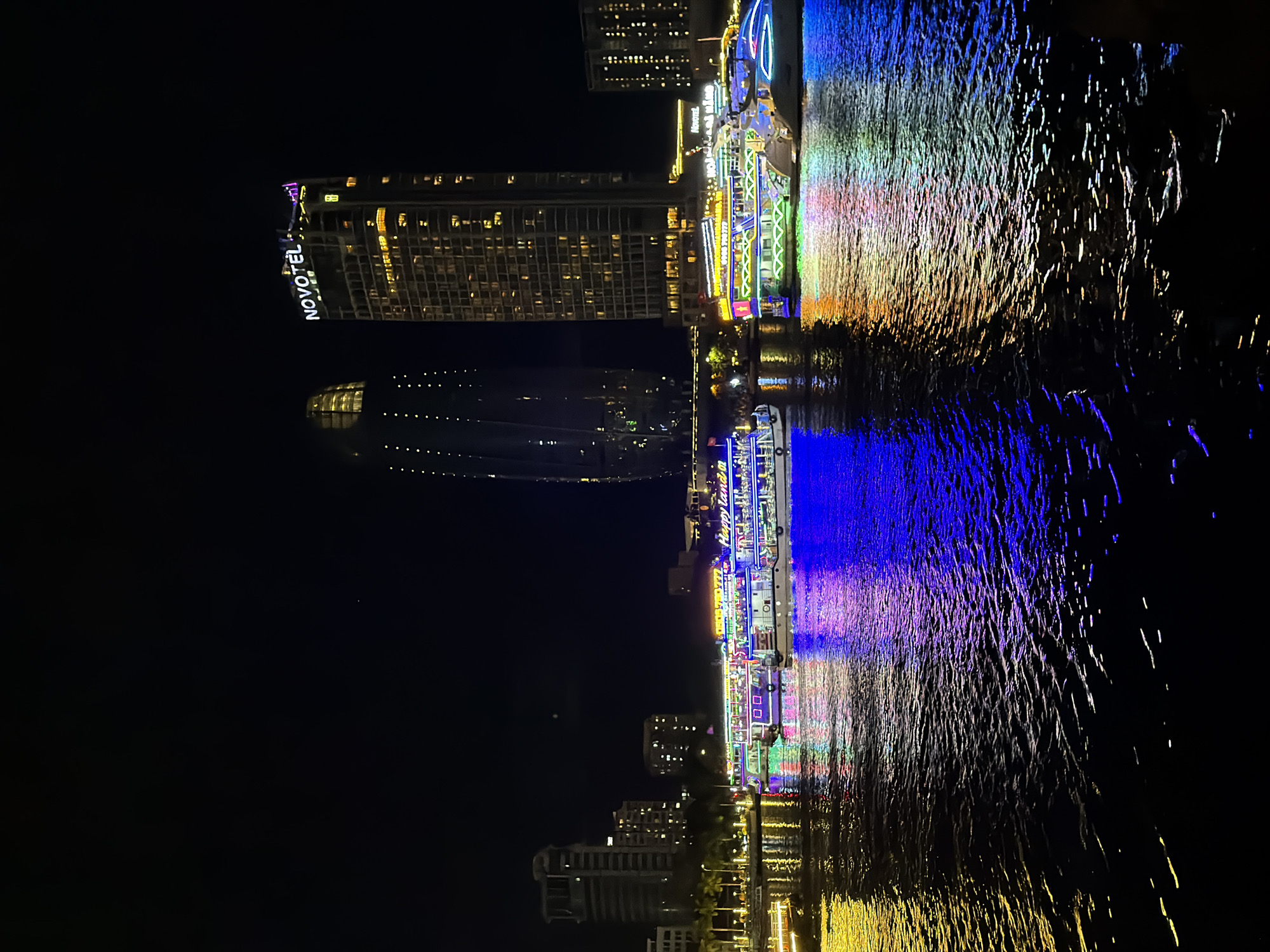 This nighttime photograph depicts the Han River in Da Nang, Vietnam, showcasing a vibrant cityscape and illuminated riverboats. In the background, two prominent skyscrapers dominate the skyline. The taller one, on the right, is the Novotel Da Nang Premier Han River, its facade lit with many windows and a bright "NOVOTEL" sign at the top. To its left is another distinctive skyscraper with a curved, textured facade, also dotted with lights.
In the foreground, the dark, rippling water of the Han River reflects the colorful lights from several large riverboats. These boats are adorned with numerous bright lights in various colors, including blue, purple, yellow, and green, creating dazzling reflections on the water's surface. Some of the boats appear to be tour vessels, with "Happy Land" visible on one of them. The riverbank on the left is lined with palm trees and other buildings, also softly illuminated. The overall scene is one of lively urban nighttime ambiance, with the illuminated architecture and colorful boat lights creating a festive atmosphere. No people are clearly discernible in the image.