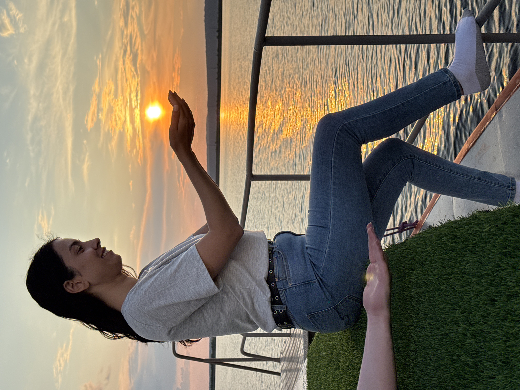A young woman with long, dark hair is sitting on the deck of a boat, facing right, with her hands cupped in front of her. The setting sun is visible in the sky, appearing as a bright orb between her hands, casting a warm glow on the water. She is wearing a light-colored t-shirt, blue jeans, a black belt, and white socks. Another person's hand is visible on the left, resting on the artificial turf-covered edge of the boat. The boat is on a body of water, with a railing in the foreground and a distant shoreline. The sky is filled with soft clouds illuminated by the sunset. The image is taken in Chaikovsky, Russia.