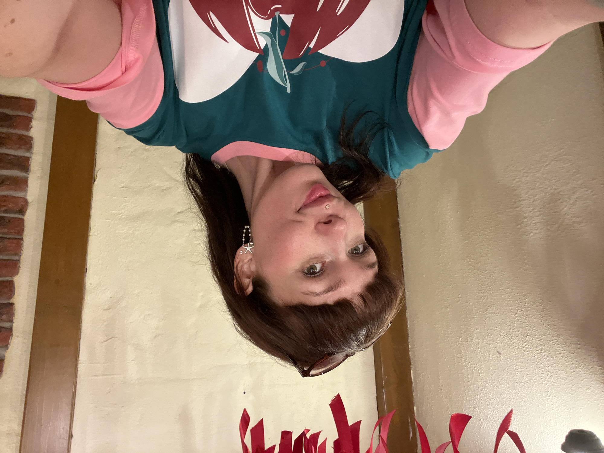 A young woman with brown hair and bangs, wearing a teal and pink long-sleeved shirt with a large red apple graphic, is taking a selfie. She has light-colored eyes, a mole on her upper lip, and star-shaped earrings. A pair of sunglasses rests on top of her head. She is indoors, with textured beige walls and wooden accents visible in the background. Red ribbons are draped across the top of the frame, suggesting a festive decoration. The image does not contain any information that places the location in Chaikovsky, Russia.