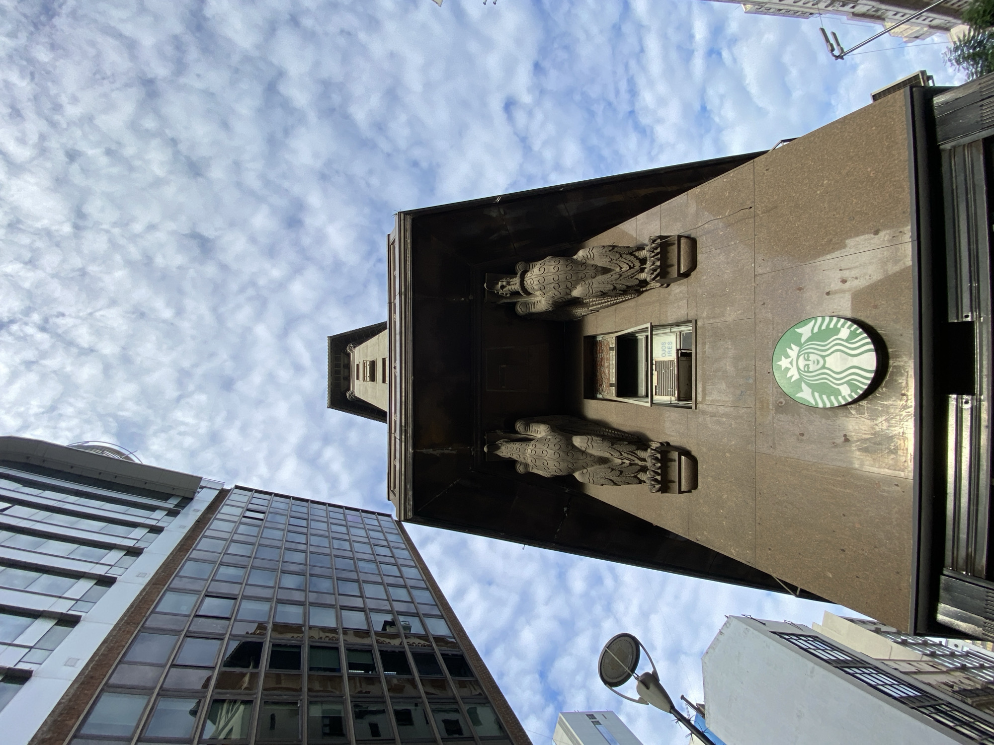 The image shows a low-angle view of a building in Buenos Aires, Argentina, featuring a Starbucks sign on its façade. The building has two stylized eagle sculptures flanking a window. The sky is visible with scattered clouds. Several other buildings are visible in the background. There are no people shown.