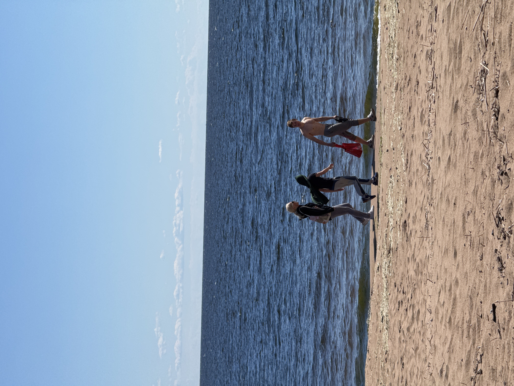 The image shows three people on a beach next to a body of water. The scene is set on a sandy beach with a clear blue sky above. The water is dark blue and appears to be choppy.
The subjects are three individuals, two appearing to be male and one of indeterminate gender from the visible portions. The person closest to the camera is wearing a dark hooded sweatshirt and dark pants, with their arms bent and hands visible. Behind them, another person is wearing a dark shirt and lighter pants, with their arm extended. The furthest person is shirtless, wearing grey shorts and is holding a red object in their hand. They are all walking along the edge of the water.
The time of day appears to be daytime, likely sunny given the clear sky and the shadows cast. The weather seems pleasant for outdoor activities. The location is suggested by the prompt to be Zelenaya Roshcha, Russia, which implies a beach or lakeside setting. There is no visible text in the image. The overall mood appears to be casual and relaxed, consistent with a day at the beach.