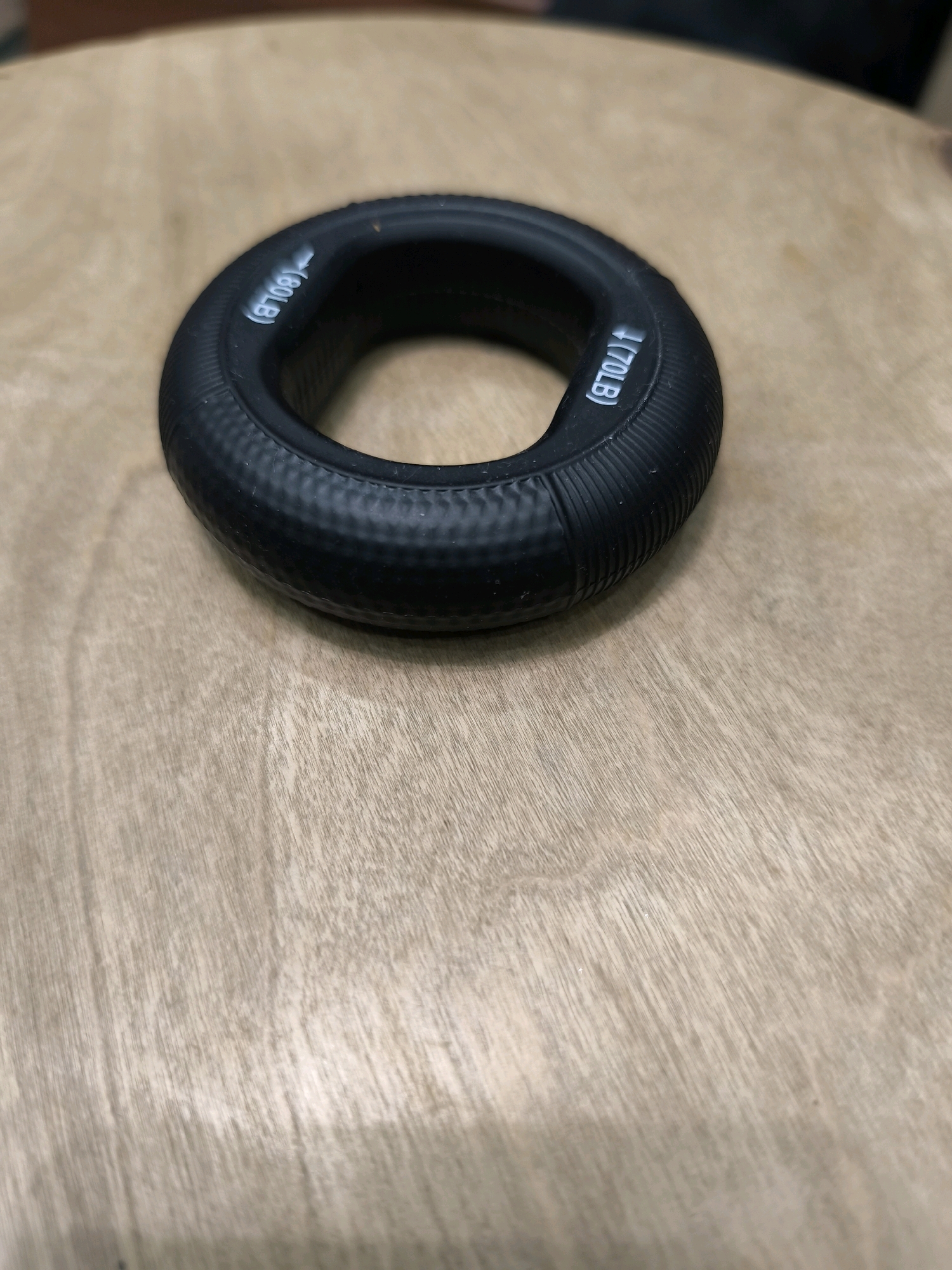 A black, circular grip strengthener lies on a light brown wooden surface. The grip strengthener has white text on its surface indicating "60LB" and "70LB". The wooden surface displays a visible wood grain pattern. The image appears to be taken from a slightly elevated angle, looking down at the grip strengthener. There is no indication of the location being Karachayevsk, Russia.