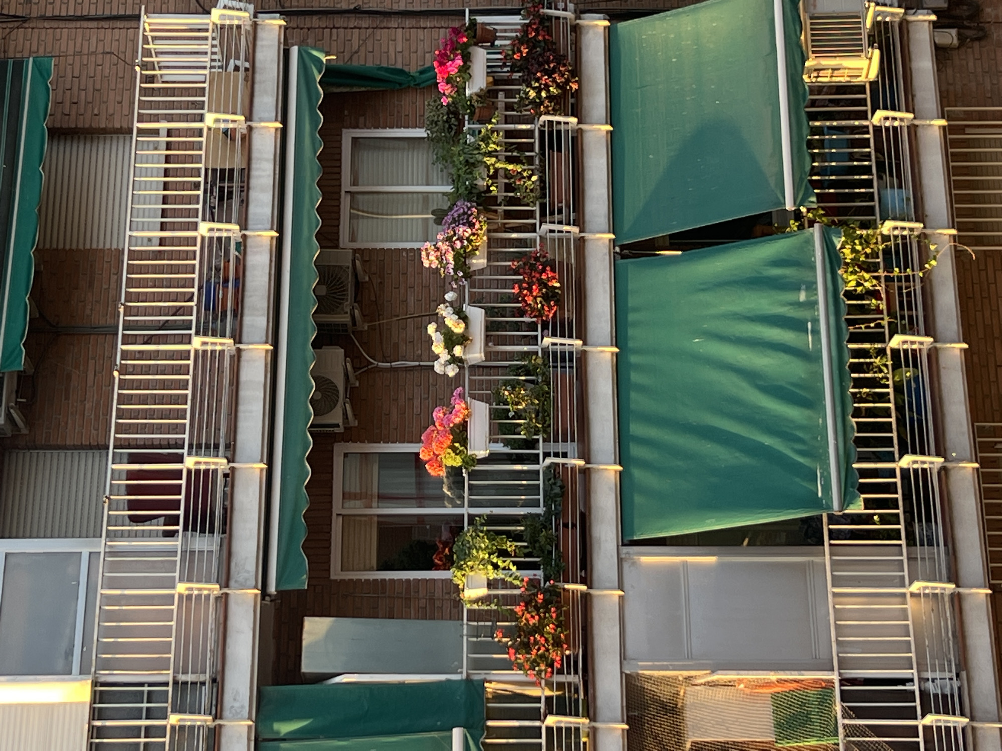The image depicts the facade of an apartment building, showcasing rows of windows and balconies adorned with potted flowers, plants, and awnings, which are characteristic features of residential buildings in Madrid, Spain. The facade is primarily constructed of brick, adding to the traditional architectural style prevalent in the city. Fire escape ladders are visible alongside the balconies. Air conditioning units are mounted on the walls below the windows. The overall scene suggests a residential neighborhood in Madrid. The warm lighting indicates the presence of sunlight, suggesting a late afternoon or early morning time frame.