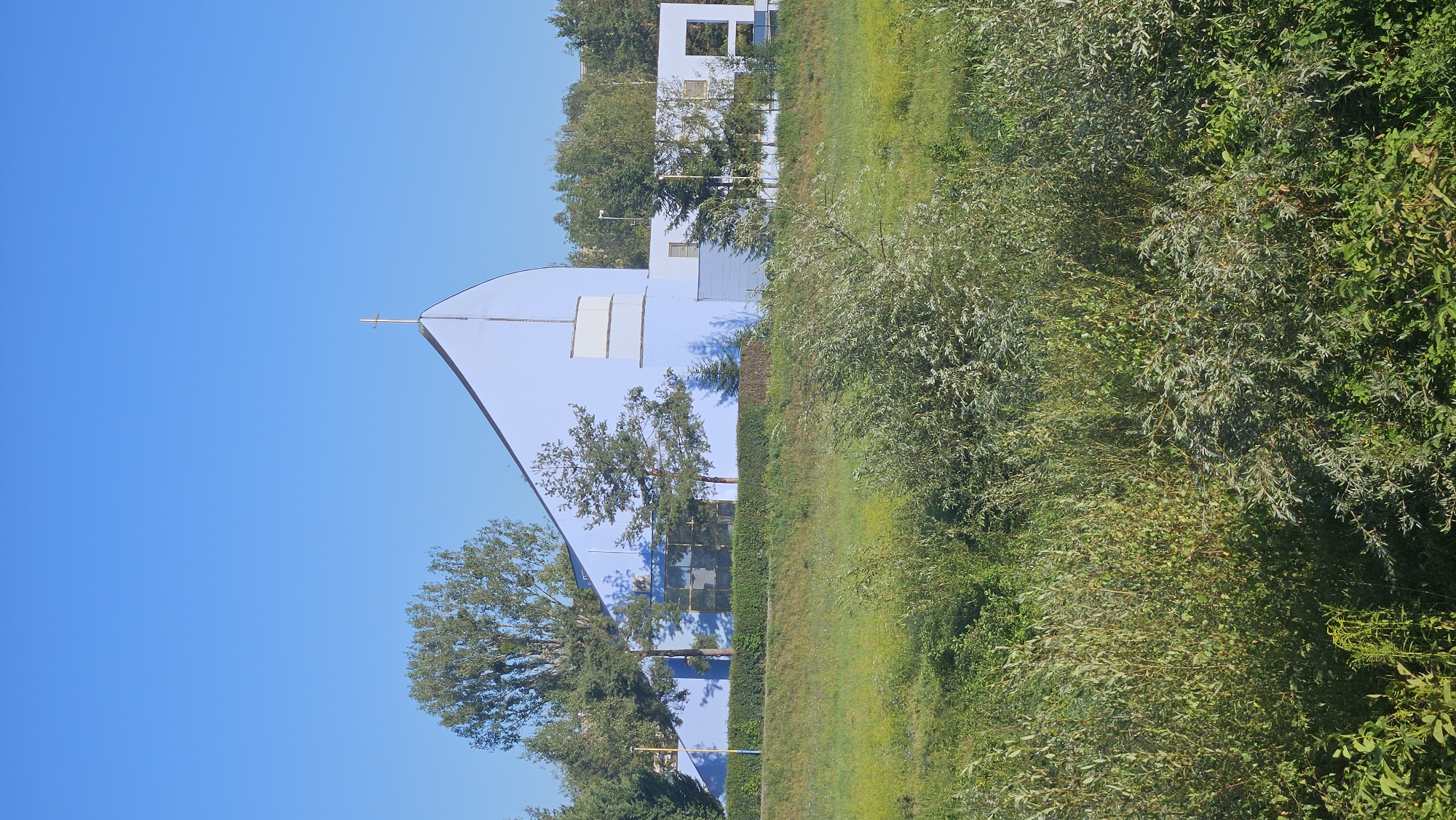 This image captures a modern church building with a distinctive light blue facade and a pointed roof topped with a golden cross. The architecture is contemporary, featuring large windows and geometric elements. The church is situated amidst lush greenery, with a vibrant, verdant field in the foreground and a line of trees and shrubs behind the building, forming a natural backdrop. The sky is a clear, bright blue, suggesting a sunny day. The scene appears to be a quiet, serene setting, likely in a suburban or semi-rural area of Prešov, Slovakia, given the context of the request. No people are visible in the frame. The overall impression is one of peace and modern spirituality within a natural environment.