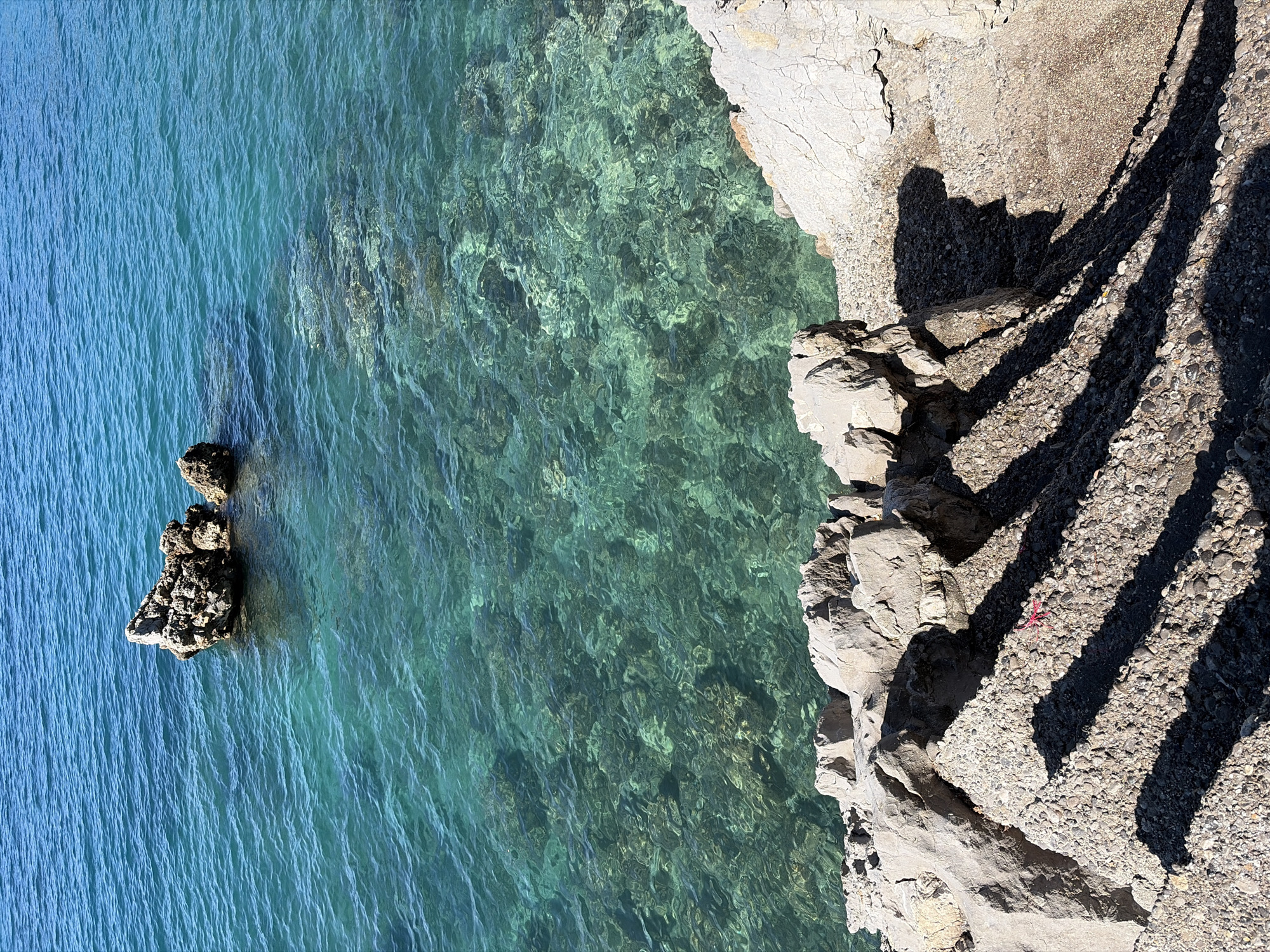 The image depicts a rocky shoreline meeting the turquoise water of the Adriatic Sea, possibly near the city of Budva, Montenegro. The perspective is a high-angle or aerial view. The shoreline consists of light-colored rocks and pebbles, casting long, dark shadows diagonally across the frame. A small, rocky islet protrudes from the sea. The water near the shore is clear, with visible seabed details, transitioning to a deeper blue further out.