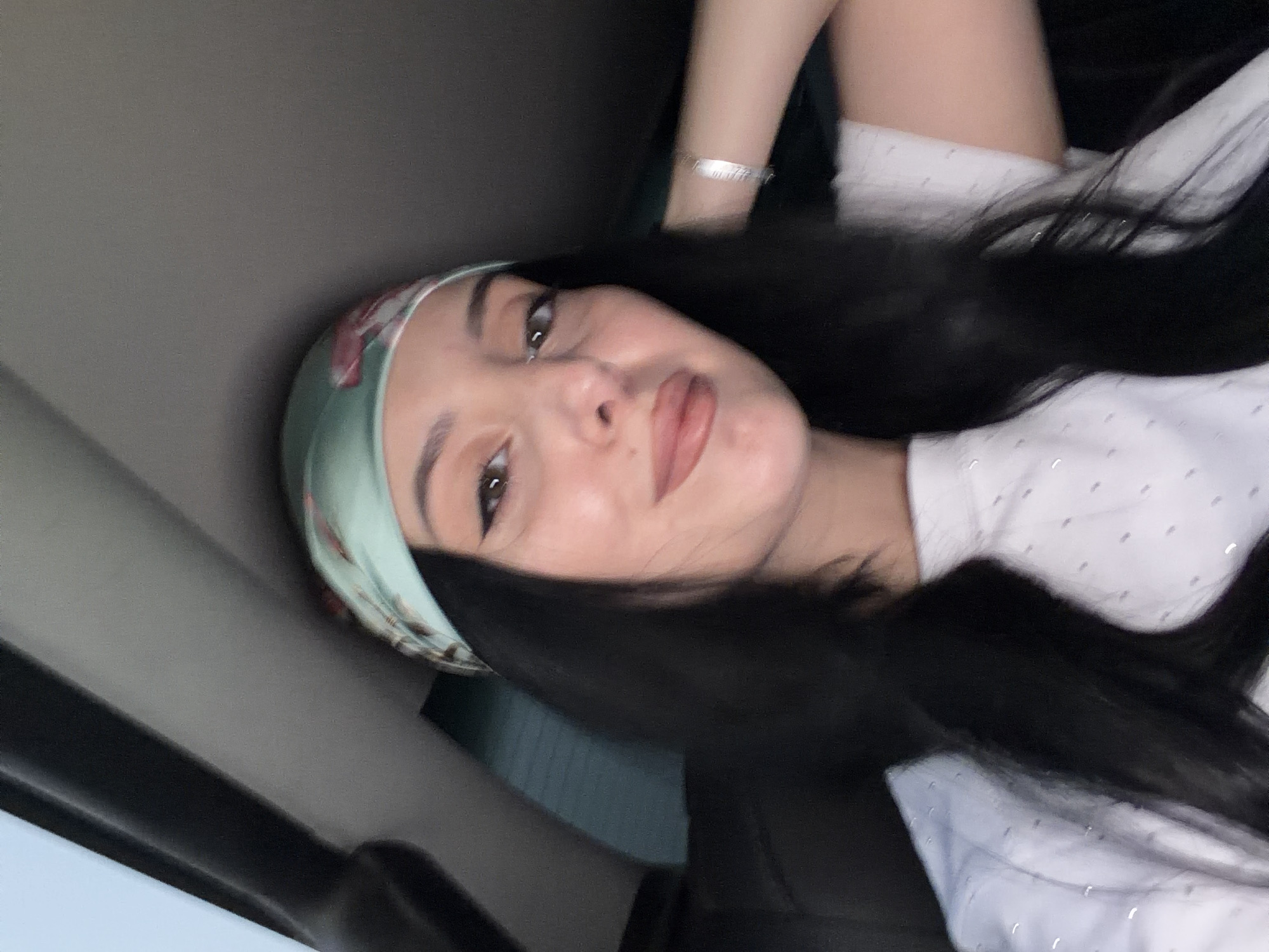A young woman with long, dark hair is visible in a selfie taken inside a vehicle. She is wearing a light blue headscarf with a floral pattern and a white t-shirt adorned with small, shimmering dots. Her makeup includes eyeliner and lipstick. A silver bracelet is visible on her right wrist. The background appears to be the interior of a car, with a dark headliner and window tinting visible. The lighting suggests it is daytime. The image does not provide any specific visual cues to indicate the location is Tbilisi, Georgia.