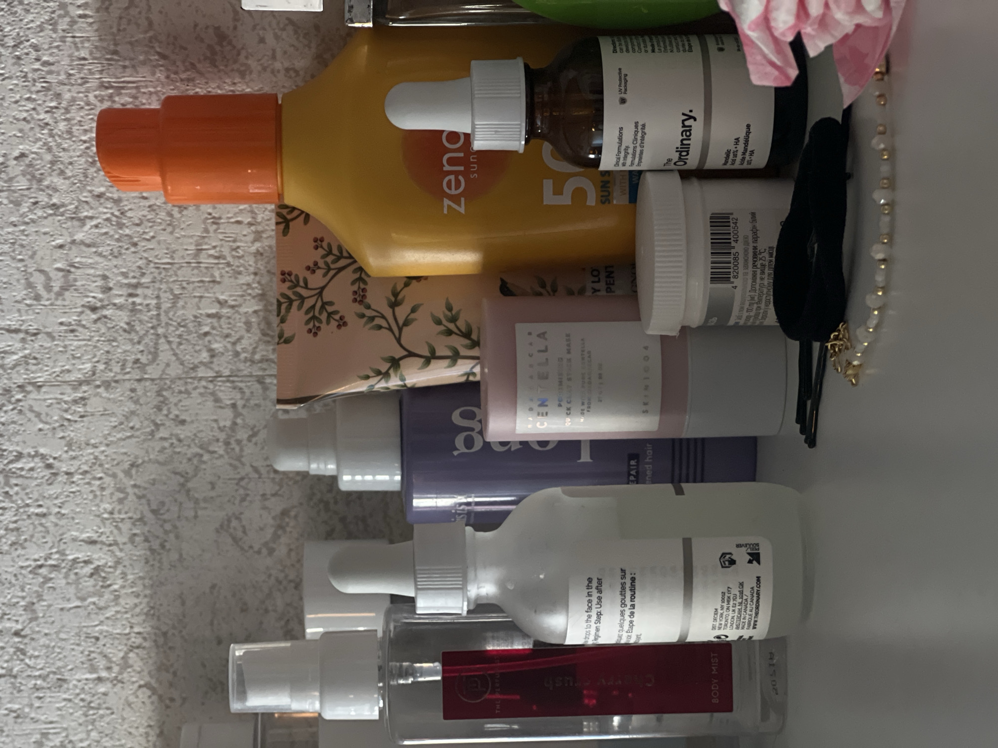 This image shows a shelf in a residential bathroom, likely in Genemuiden, Netherlands. On the shelf are various skincare and personal care products. From left to right, there is a clear spray bottle labeled "BODY MIST" with a red label, a white dropper bottle, a purple bottle, a cylindrical pink container, and a large yellow bottle with an orange cap, labeled "zendium sunscreen". There is also a small brown dropper bottle labeled "The Ordinary". Several hair accessories, including bobby pins and a beaded bracelet, are visible in the foreground. The background features a textured, light-colored wall. The lighting is soft, suggesting ambient indoor light. There are no people present in the image.