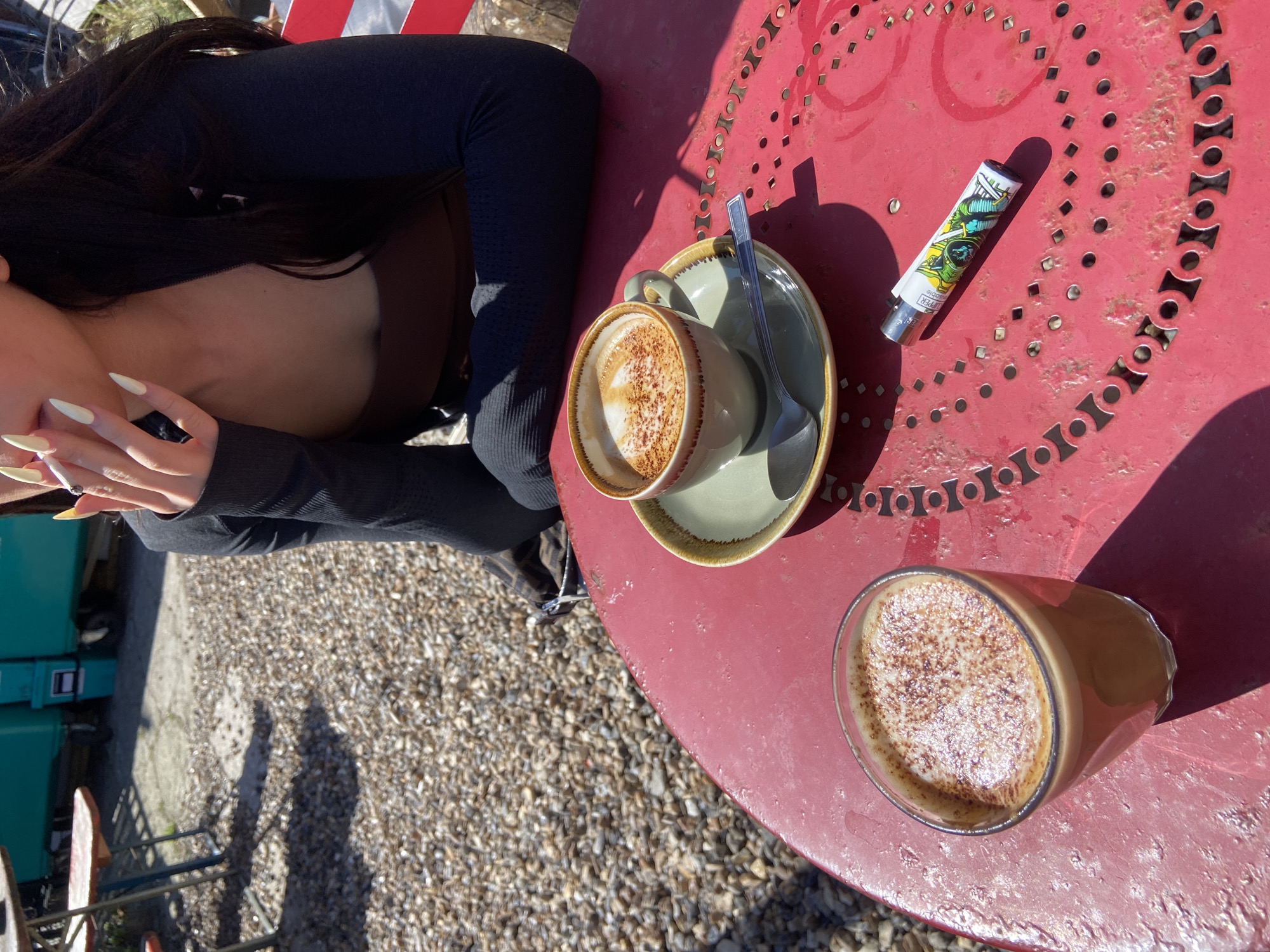 The scene is captured outdoors, likely at a café in London, United Kingdom. A woman with long dark hair and long fingernails sits at a round, red, perforated table, wearing a dark long-sleeved shirt. On the table, there are two cups of coffee or cappuccinos, one in a green cup and saucer with a spoon, and the other in a clear glass. A Clipper lighter rests near the cup and saucer. The table is situated on a gravel surface, and glimpses of other outdoor furniture and surroundings can be seen in the background.