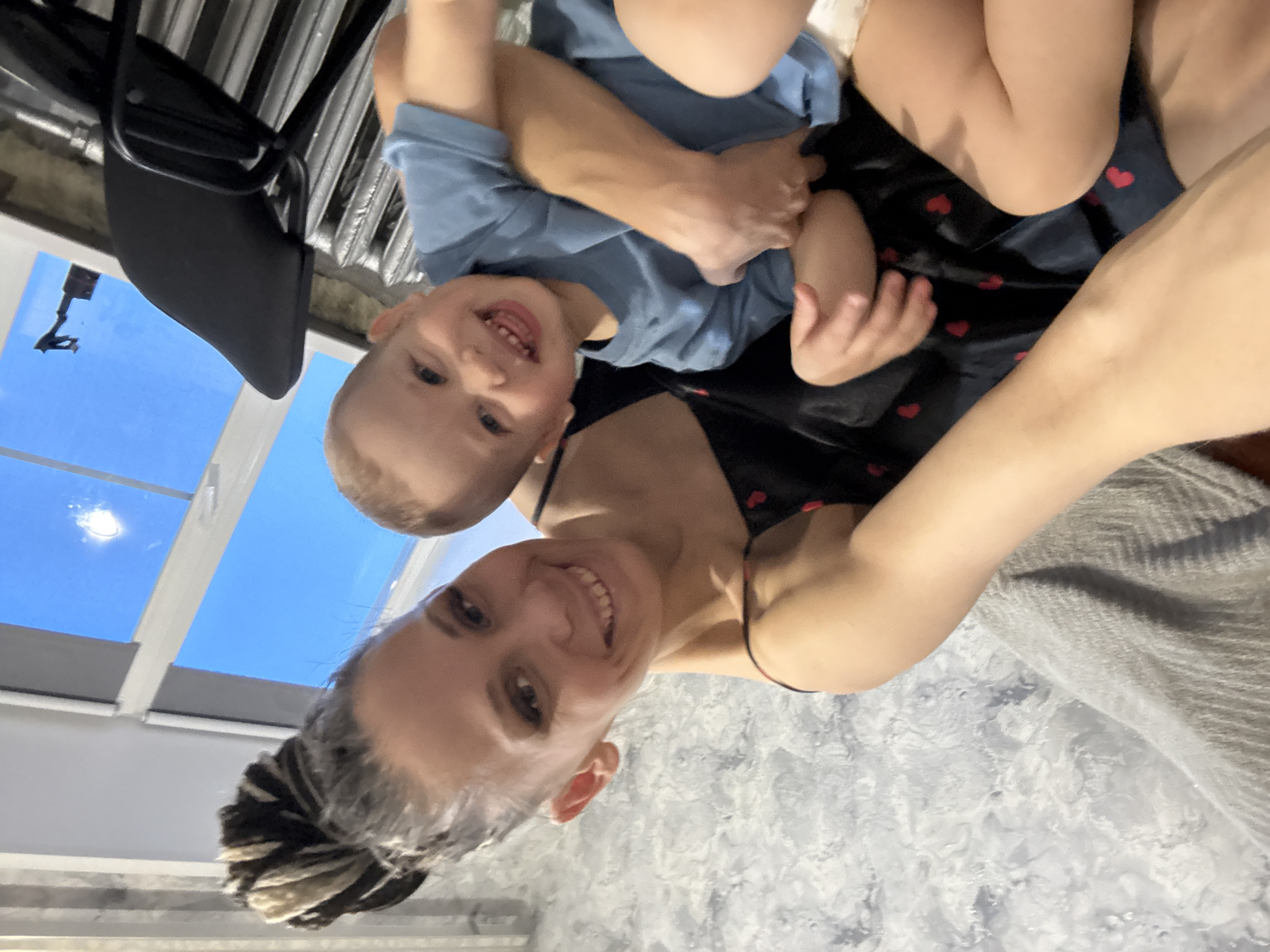 A woman and a child are in an interior. The woman is holding the child. The child is wearing a blue shirt, and the woman is wearing a black top with a heart pattern. A black chair is visible, and a window shows a blue sky. The image was likely taken indoors in an apartment or house with a textured gray wall and a patterned gray blanket. Based on the information provided, the photo might have been taken in an apartment in Rusanovo, Russia. However, specific details to confirm that location are absent from the image.