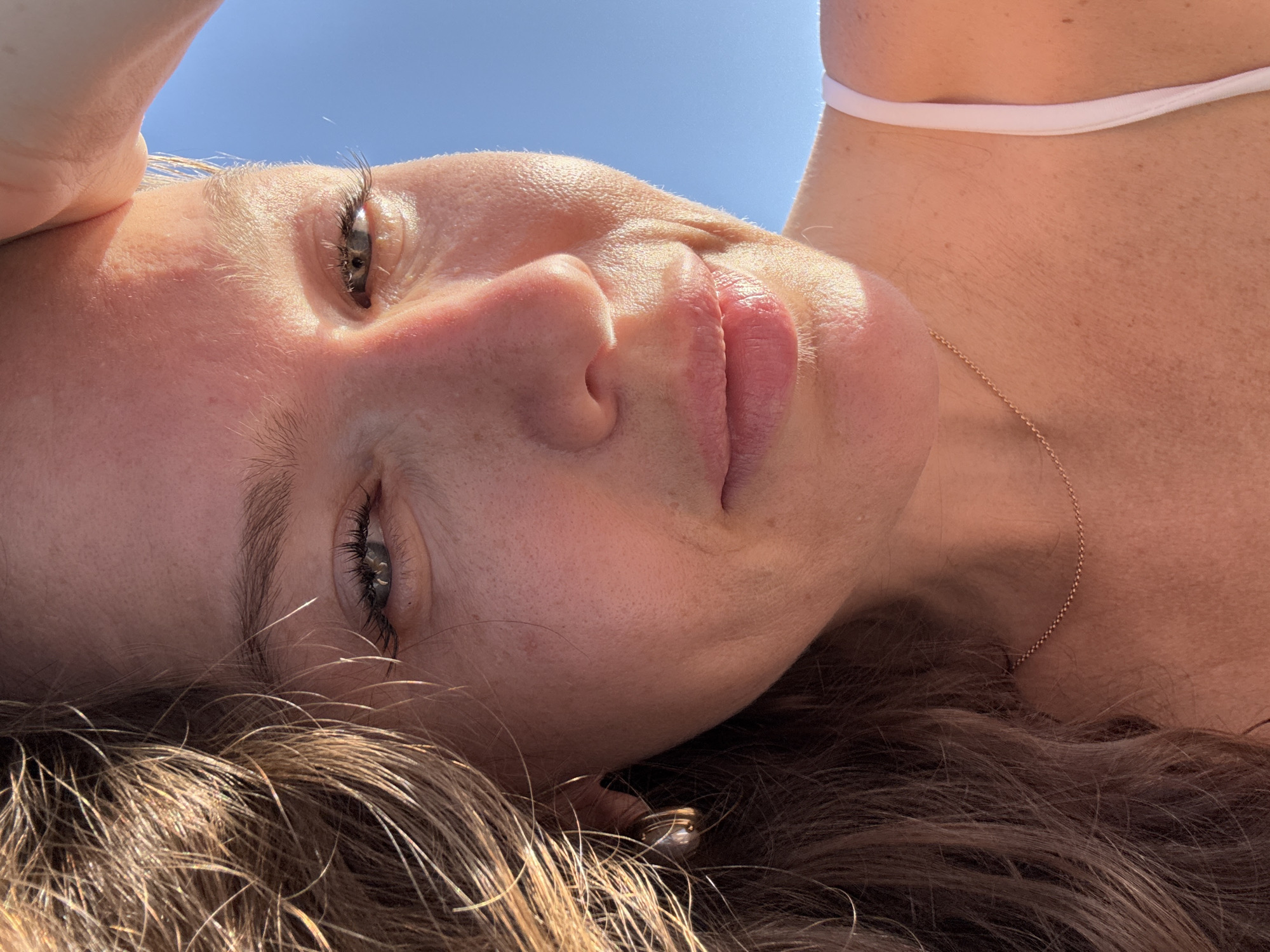 A medium shot, eye-level, close-up photograph captures a woman from the chest up, facing the camera with a slight smile. Her head is tilted to the left, with her right hand raised to her forehead, shielding her eyes from the sun. She has light brown hair, styled loosely, with strands falling across her face. Her eyes are light green, and she has a smattering of freckles across her nose and cheeks. She is wearing a delicate gold chain necklace and a white bikini top. The background is a clear, bright blue sky. The lighting suggests it is daytime, with the sun casting highlights on her skin and hair. The overall impression is one of a casual, outdoor portrait, likely taken on a sunny day in Dubai, United Arab Emirates, perhaps at a beach or poolside location.