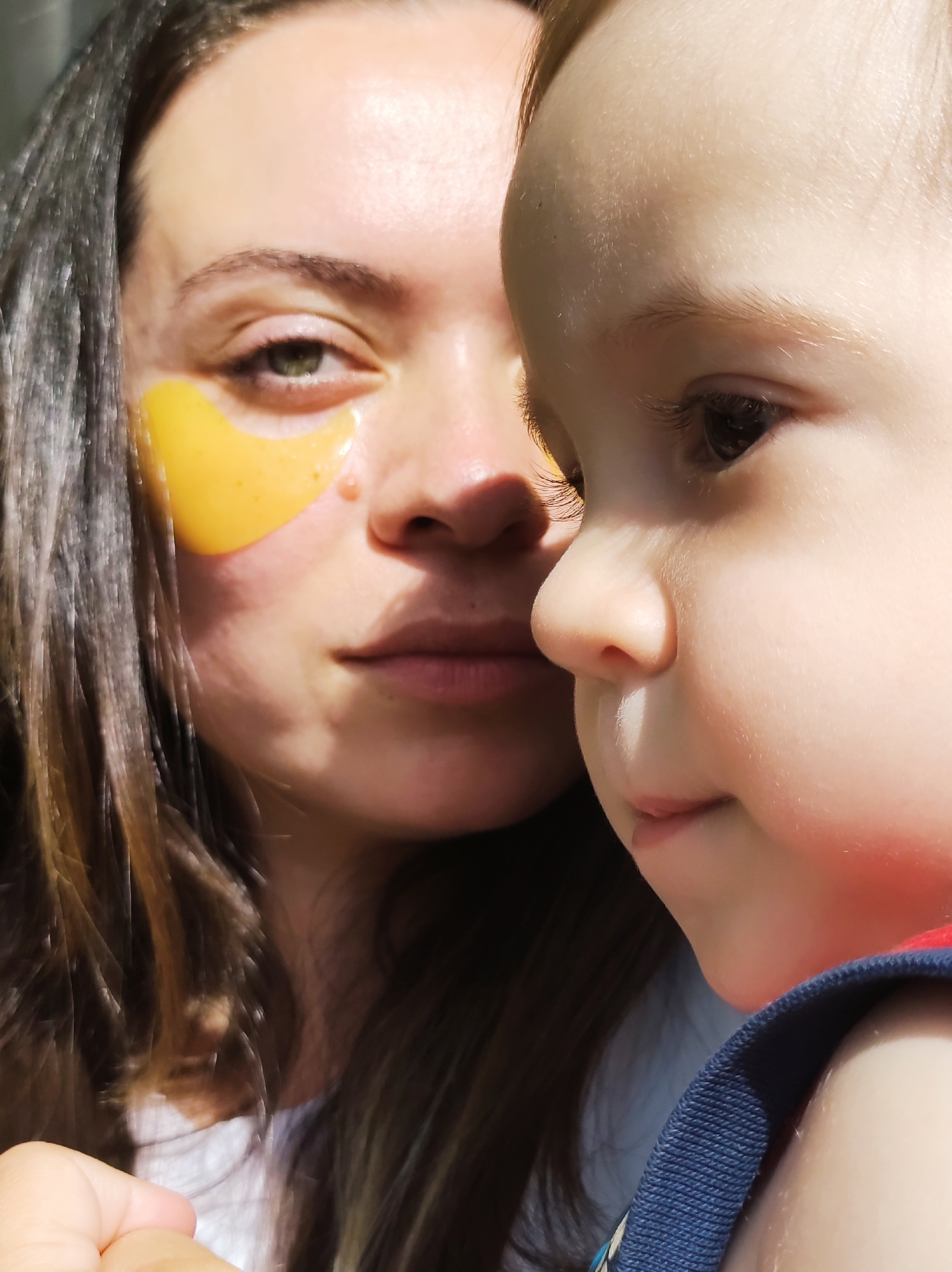 The image presents a close-up of a woman and a child, possibly in an indoor setting. The woman has dark hair and is wearing a gold-colored eye mask under one eye. Her facial expression is neutral. The child, positioned in the foreground and slightly to the right, has fair skin and hair. The child's face is partially in focus, showing a profile view. The clothing worn by the child includes a blue and possibly red garment. There is no visible indication of Chaikovsky, Russia, or any specific location in the scene. The interaction appears to be a close, familial moment.