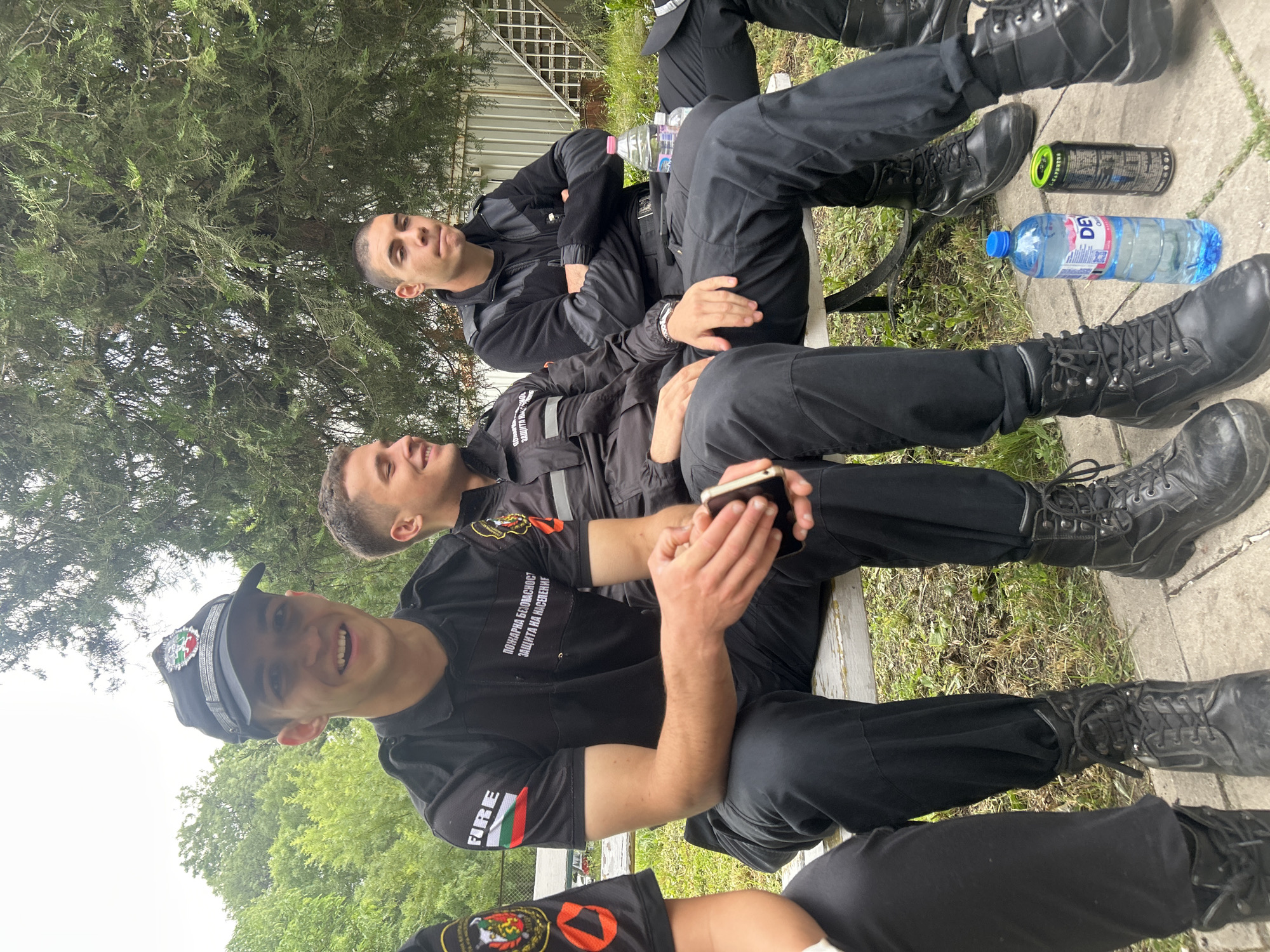 Three young men wearing black uniforms and boots are sitting on a concrete bench in an outdoor setting, likely in Sofia, Bulgaria, judging by the text on their uniforms. The men appear to be taking a break. One man in the front is smiling towards the camera, holding a phone. The man behind him is looking off to the side and the third man is looking up. A bottle of water and a can are on the ground near their feet. The area around the bench is grassy and there are trees in the background. A fence is partially visible in the background.
