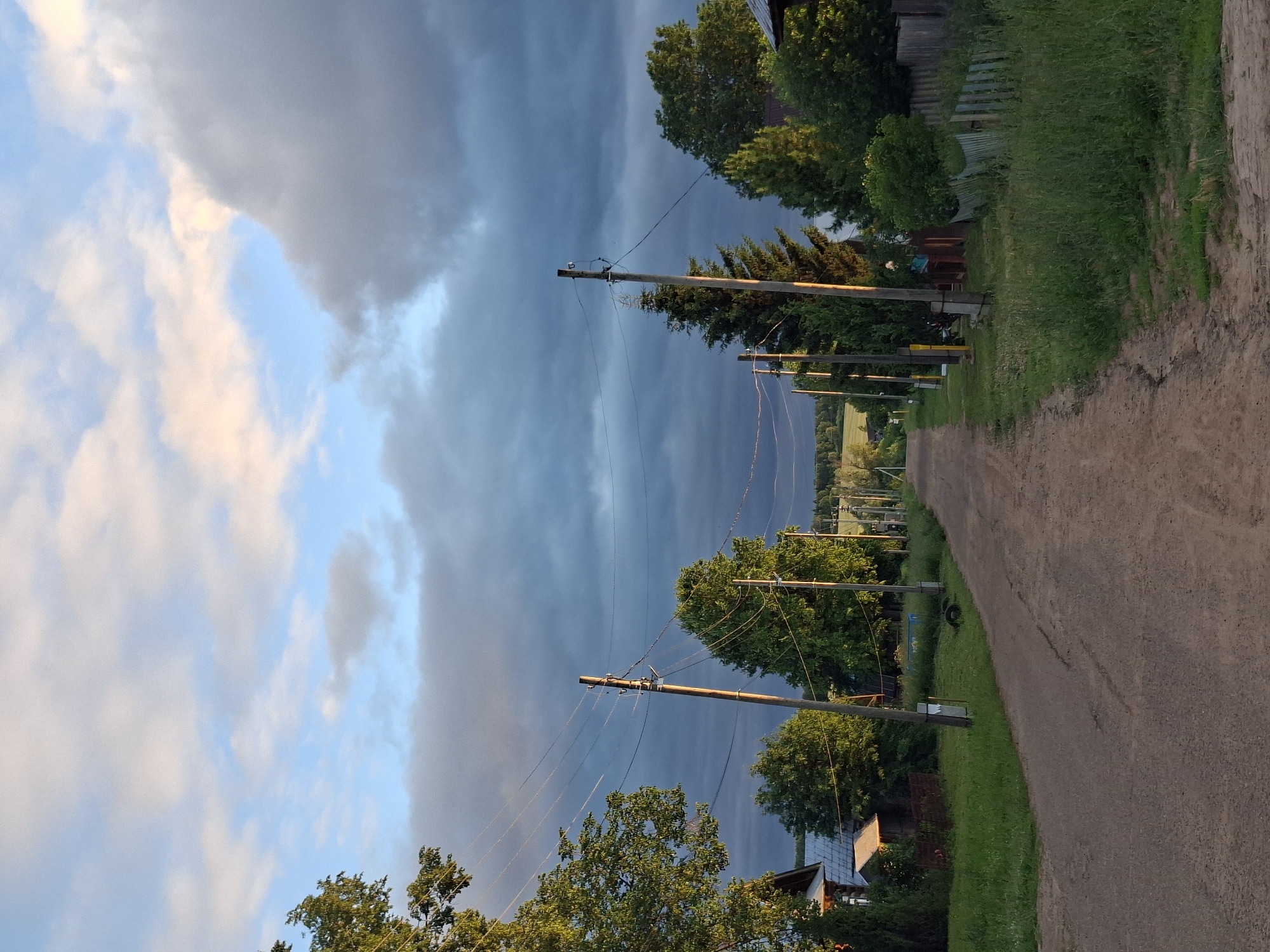 This image depicts a rural road in Karsha, Russia, with a dramatic sky filled with dark storm clouds and streaks of lighter, sunlit clouds. The time of day appears to be late afternoon or early evening, as indicated by the warm light on the clouds and the shadows cast by the trees. The road itself is unpaved, likely a dirt or gravel path, and curves to the right, disappearing into the distance.
Along the left side of the road, a line of wooden utility poles with power lines stretches into the distance. These poles are interspersed with lush green trees, some of which are tall and mature, while others are smaller bushes. Beyond the trees, glimpses of houses and fences can be seen, suggesting a small village or rural settlement. The right side of the road is bordered by a grassy verge and more trees.
The weather is a prominent element of the scene, with the contrast between the dark, heavy storm clouds and the illuminated parts of the sky creating a sense of anticipation or even foreboding. The presence of the utility poles and houses confirms a human-inhabited landscape, but the overall mood is dominated by the natural spectacle of the approaching storm. No people are visible in the frame. There is no visible text in the image.