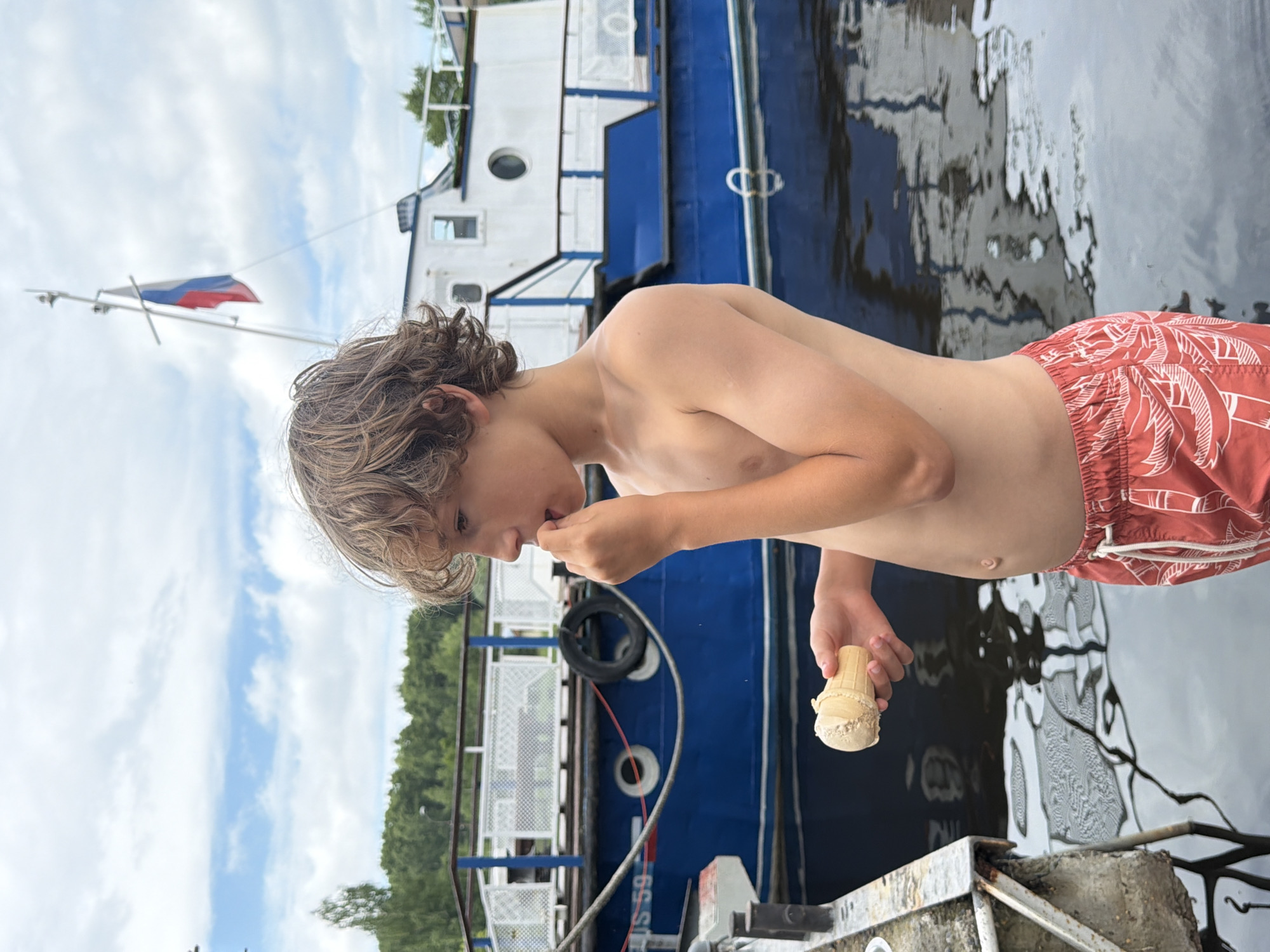 This is a vertical, medium shot photograph taken outdoors. The main subject is a young boy with light brown, curly hair, wearing red swim trunks with a white palm leaf pattern. He is positioned in the foreground, leaning forward and eating an ice cream cone. The boy's face is partially obscured as he focuses on his treat. His torso and arms are visible, showing smooth skin.
Behind the boy, a blue and white boat is docked. The boat has several windows and a white railing. The Russian flag is visible flying from a flagpole on the boat, providing a strong clue to the location. The water next to the boat is dark and reflects the sky and the boat, creating a textured surface. In the background, there is a line of green trees under a sky filled with white, wispy clouds.
The activity taking place is the boy enjoying an ice cream cone, likely on a warm day given the presence of swim trunks and the outdoor setting. The overall mood seems relaxed and casual, suggesting a leisurely moment by the water. There is some visible text on the side of the boat, which appears to be a registration number: "US 59". The lighting suggests it is daytime, likely with overcast or partly cloudy conditions.