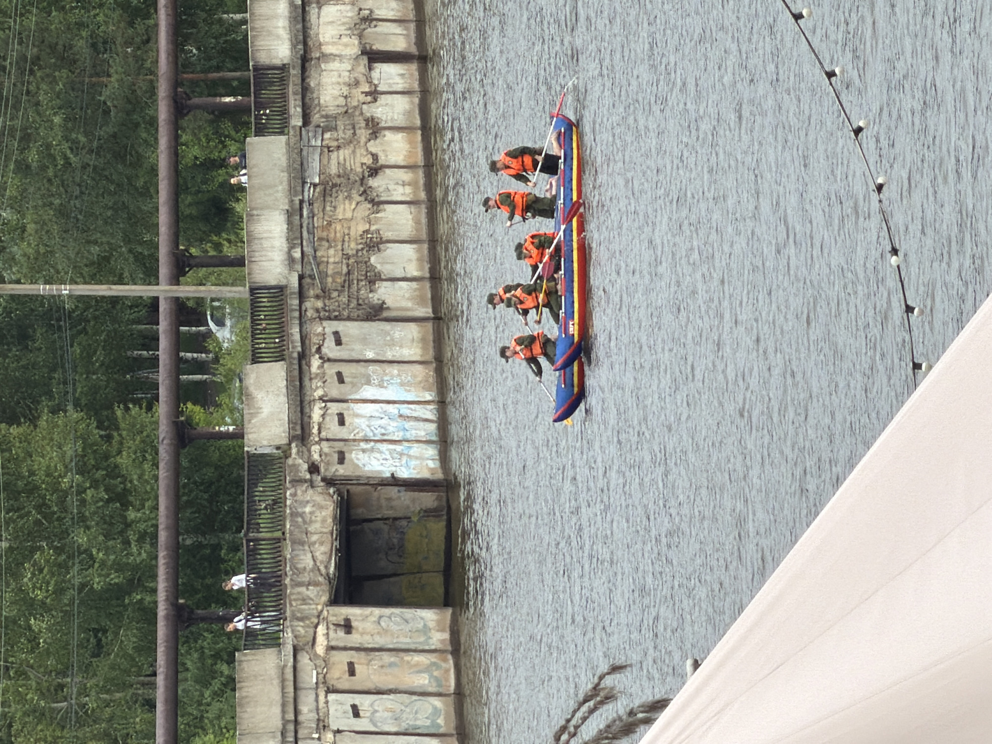 The image captures a body of water with a raft in the foreground, carrying five individuals wearing orange life vests and dark caps. They are actively paddling the raft. In the background, a concrete structure with graffiti lines the water's edge, and beyond that, a dense green forest is visible, with a large pipe supported by pillars running horizontally across the frame. Two individuals, one in a white top and the other in a dark top, are standing on a railing above the concrete structure. A white tent-like structure is partially visible in the lower left corner of the image. The overall scene suggests an outdoor recreational or training activity on the water, with industrial and natural elements in the background. This image is located in the city of Votkinsk, Russia.