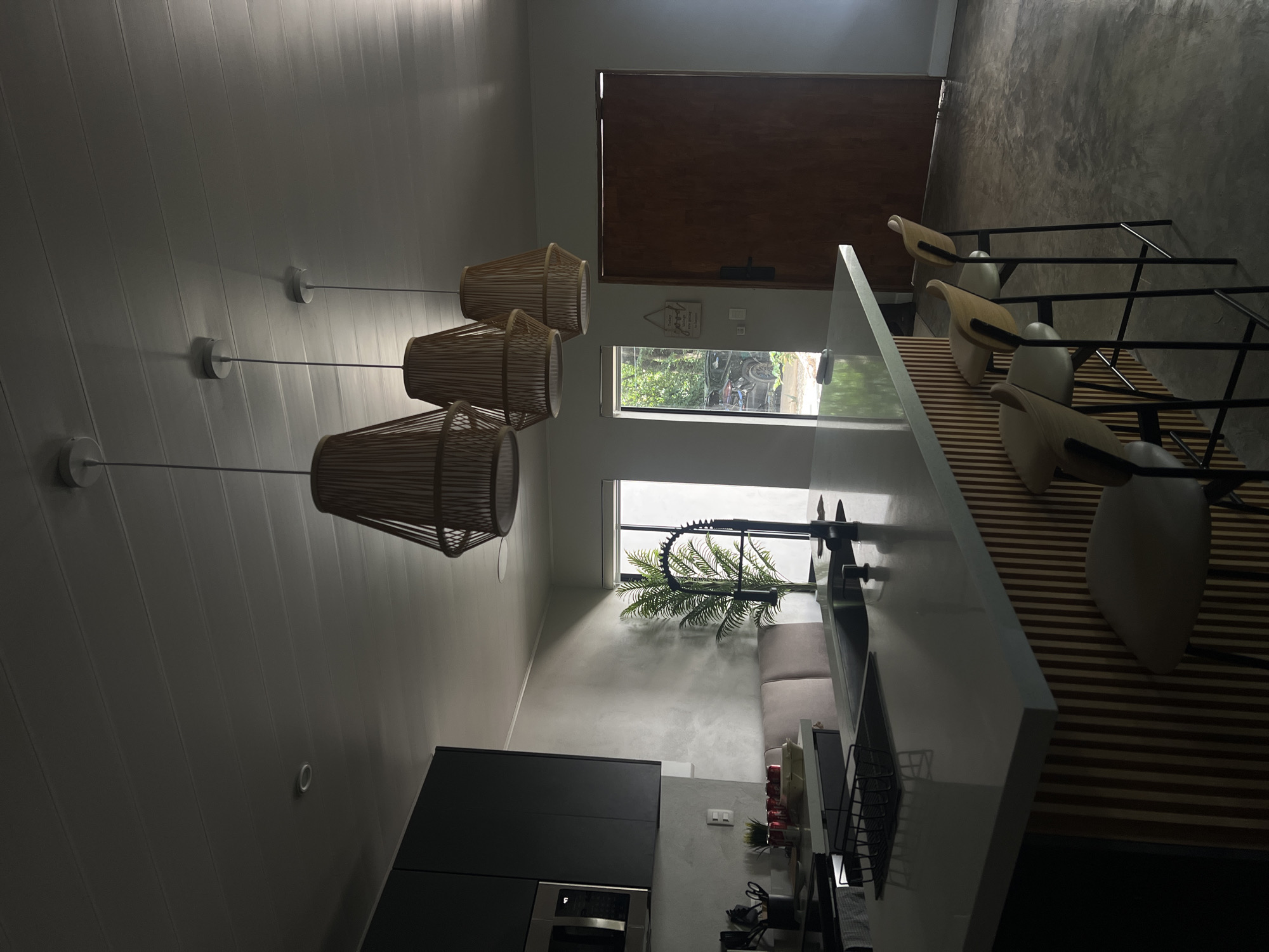 The image depicts an interior scene of a modern kitchen and dining area. The focal point is a large kitchen island with a concrete countertop, featuring a black faucet and sink. Three pendant lights with woven rattan shades hang above the island. The island's base is constructed with vertical wooden slats. Three modern bar stools with light-colored seats and wooden backs are positioned at the island. To the left, dark cabinets and a microwave are visible. A large potted plant stands near a window that looks out onto a green, leafy exterior, with a vehicle partially visible. To the right, a tall wooden door is present. The floor appears to be polished concrete. The overall lighting is dim, with natural light entering from the window. The scene is clean and uncluttered, suggesting a residential setting in Pinilla, Costa Rica. No people are visible in the image.
