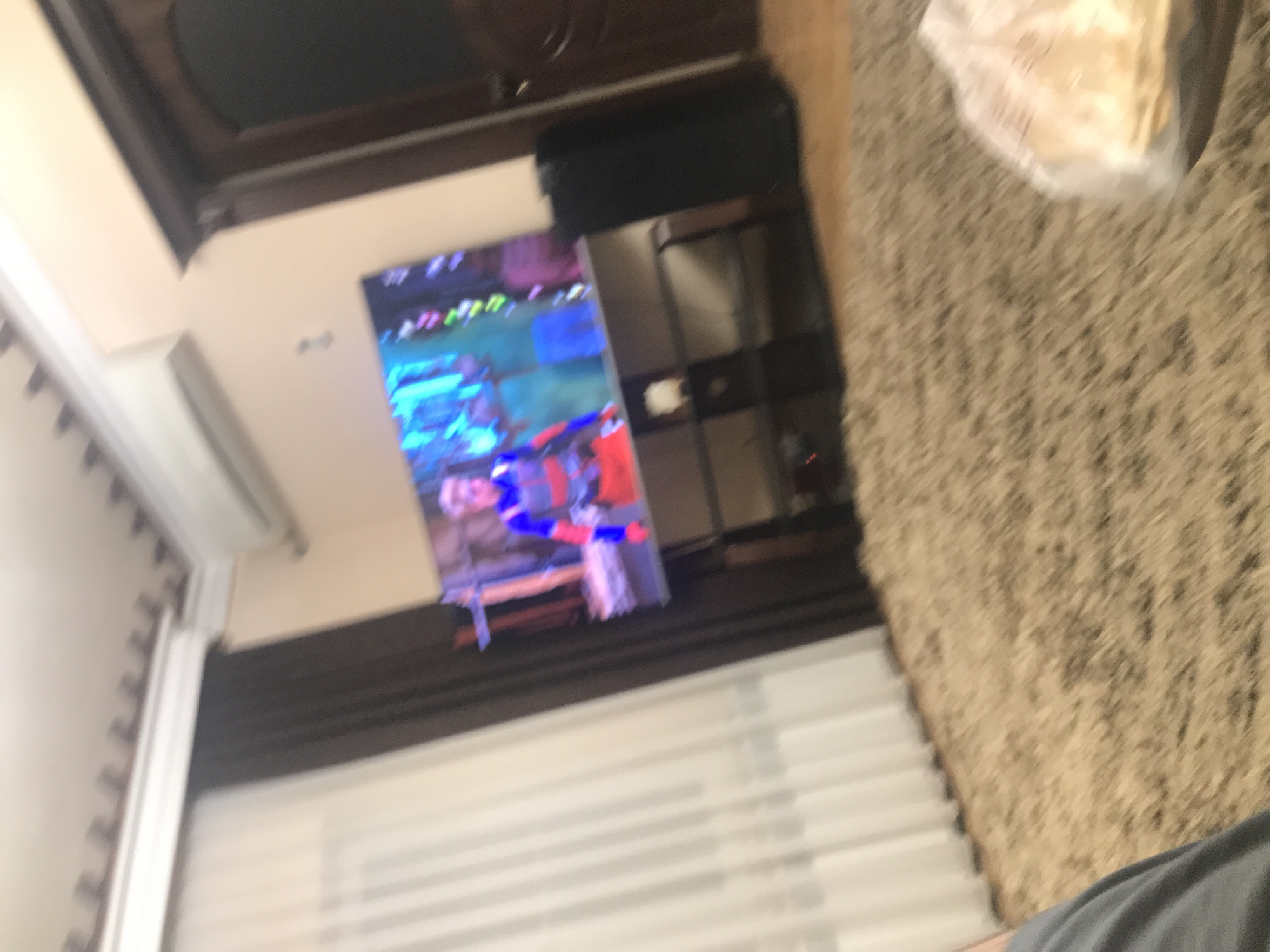 The image shows a blurred indoor scene. A television screen displays a figure in red, white, and blue, set against a background of green and blue. A shelf-like unit stands to the right of the TV. The scene also includes a white window shade, a beige rug, and a glimpse of a ceiling and a wall. Given the image's context, without external references or information, it's not possible to pinpoint the exact location in Yerevan, Armenia.