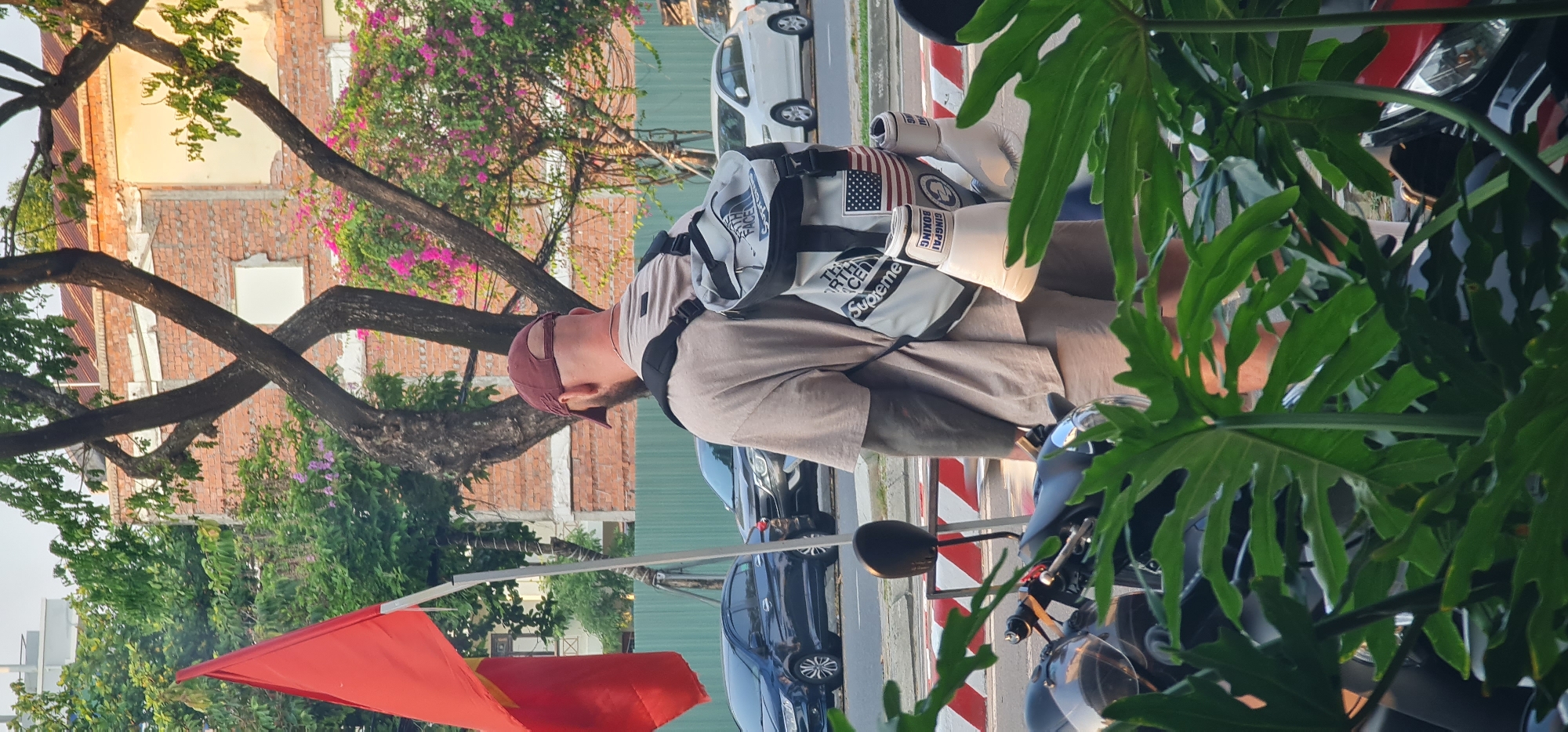 A medium shot shows a man standing next to a motorcycle, likely in Da Nang, Vietnam, near a street corner marked by red and white stripes, with a red flag pole visible in the lower left corner. The man is wearing a maroon baseball cap, a tan T-shirt, and a backpack with "THE NORTH FACE" and "Supreme" branding. He has boxing gloves attached to the backpack. The background includes a partially dilapidated brick building with colorful flowering trees, suggesting an urban environment. Vehicles are parked along the street, and there is dense green foliage in the foreground partially obscuring the view.