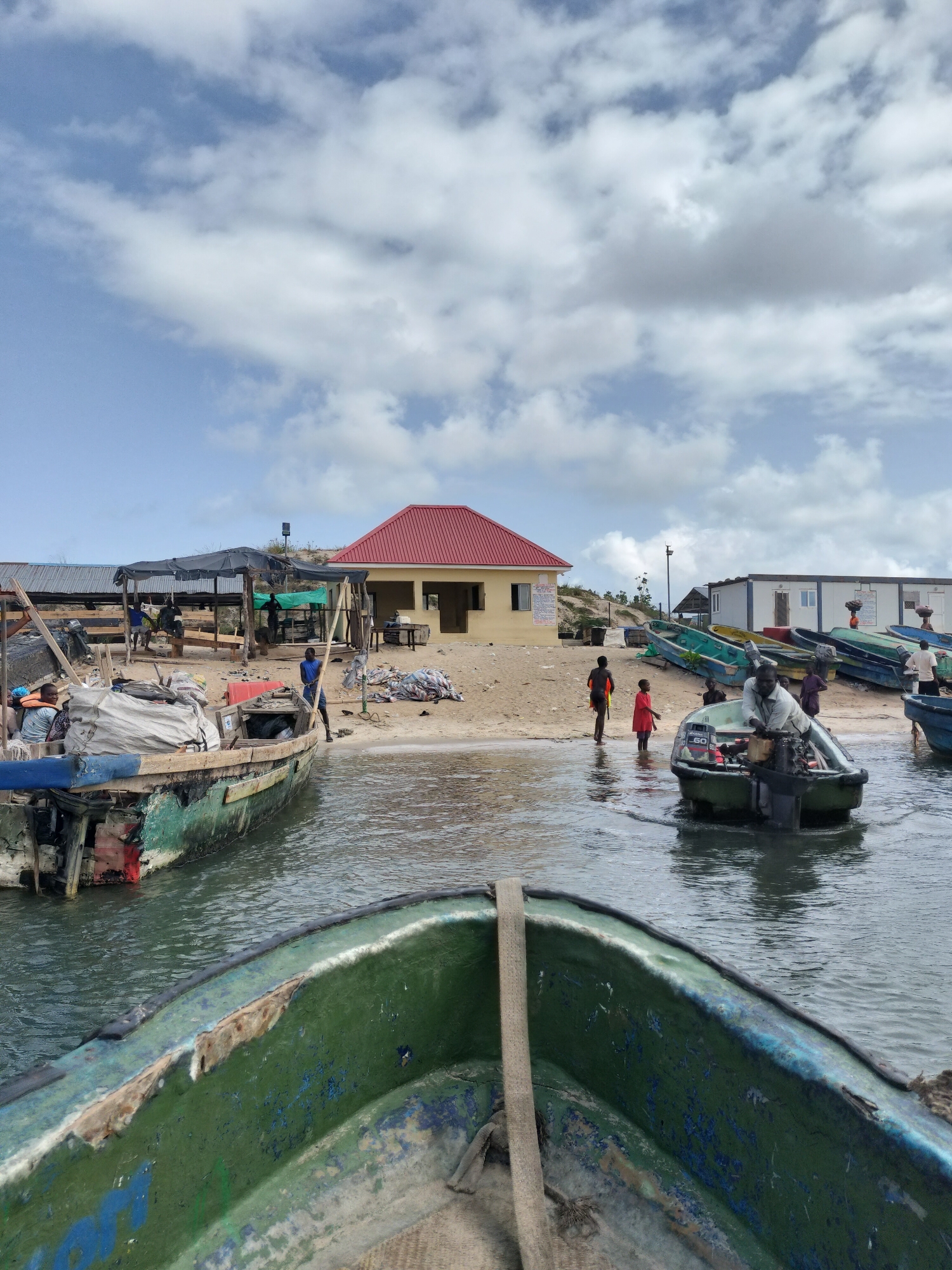 The image depicts a waterfront scene in a fishing community, likely in Lagos, Nigeria. Several small, weathered fishing boats are visible, some in the water and others pulled onto the sandy shore. One boat, in the foreground, appears to be the vantage point of the photo. The water is calm. On the shore, there is a variety of activity. Some people are gathered near the boats, possibly preparing for fishing or unloading cargo. Two children stand in the water. A man stands in a boat with a 60hp engine. Buildings and other structures are located behind the beach. The sky is overcast with white clouds.