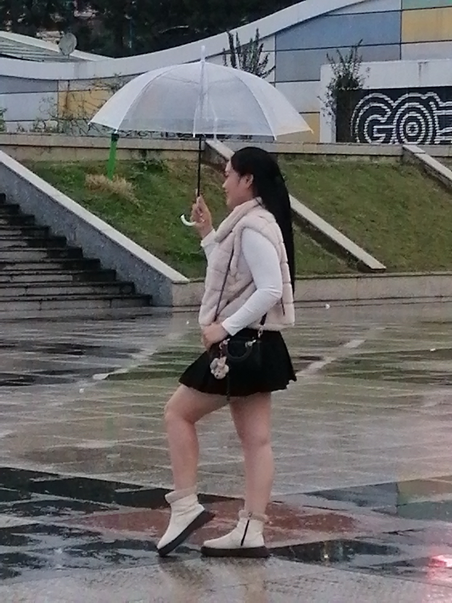 The image captures a woman holding a transparent umbrella while walking on wet, patterned pavement. She is wearing a light-colored puffer vest over a long-sleeved white shirt, a black mini-skirt, and light-colored ankle boots with fur trim. A small black purse with a flower charm hangs from her shoulder. Behind her, a grassy slope leads up to a building with a colorful, geometric facade and a distinctive "GO" graphic. Stairs are visible to the left of the frame. The overall scene suggests an outdoor urban setting in Da Lat, Vietnam, possibly on a rainy or recently rained-on day, given the wet pavement and umbrella.