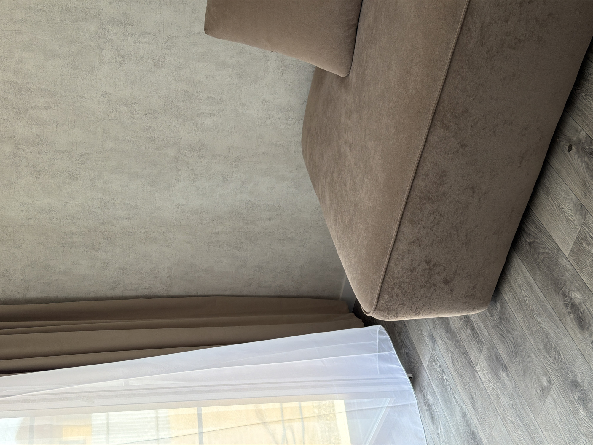 A low-angle shot captures a portion of a room, likely in Chaikovsky, Russia, featuring a beige sofa with a matching pillow. The sofa occupies the upper right quadrant of the image, and its side aligns with grey wooden floorboards. Adjacent to the sofa, a window is partially visible at the bottom of the frame, covered by both a beige curtain and a sheer white curtain. The left side and upper portion of the image is dominated by a light-grey, textured wall. The scene is stationary, devoid of people or action. The environment suggests an interior residential space.