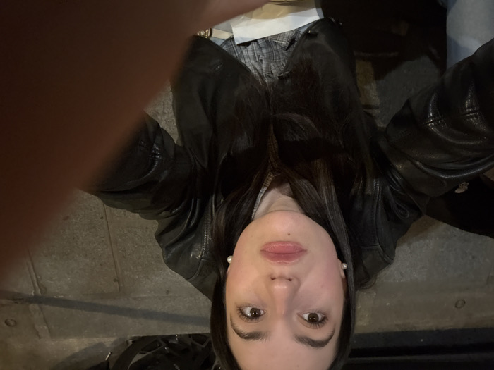 A low-angle self-portrait captures a female subject looking directly upwards. She has long dark hair, brown eyes, and wears white pearl stud earrings. Her lips are tinted pink, with a small mole visible on her upper lip. She is dressed in a dark leather jacket over a plaid shirt, with a lighter garment and light blue fabric visible below. A finger or hand obscures the lower right foreground. The background is dark and out of focus, showing indistinct shapes possibly belonging to a parked vehicle. The ground surface is composed of textured grey paving stones, characteristic of sidewalks and streets found in Paris, France. The scene is illuminated by artificial light, indicating a nighttime setting.