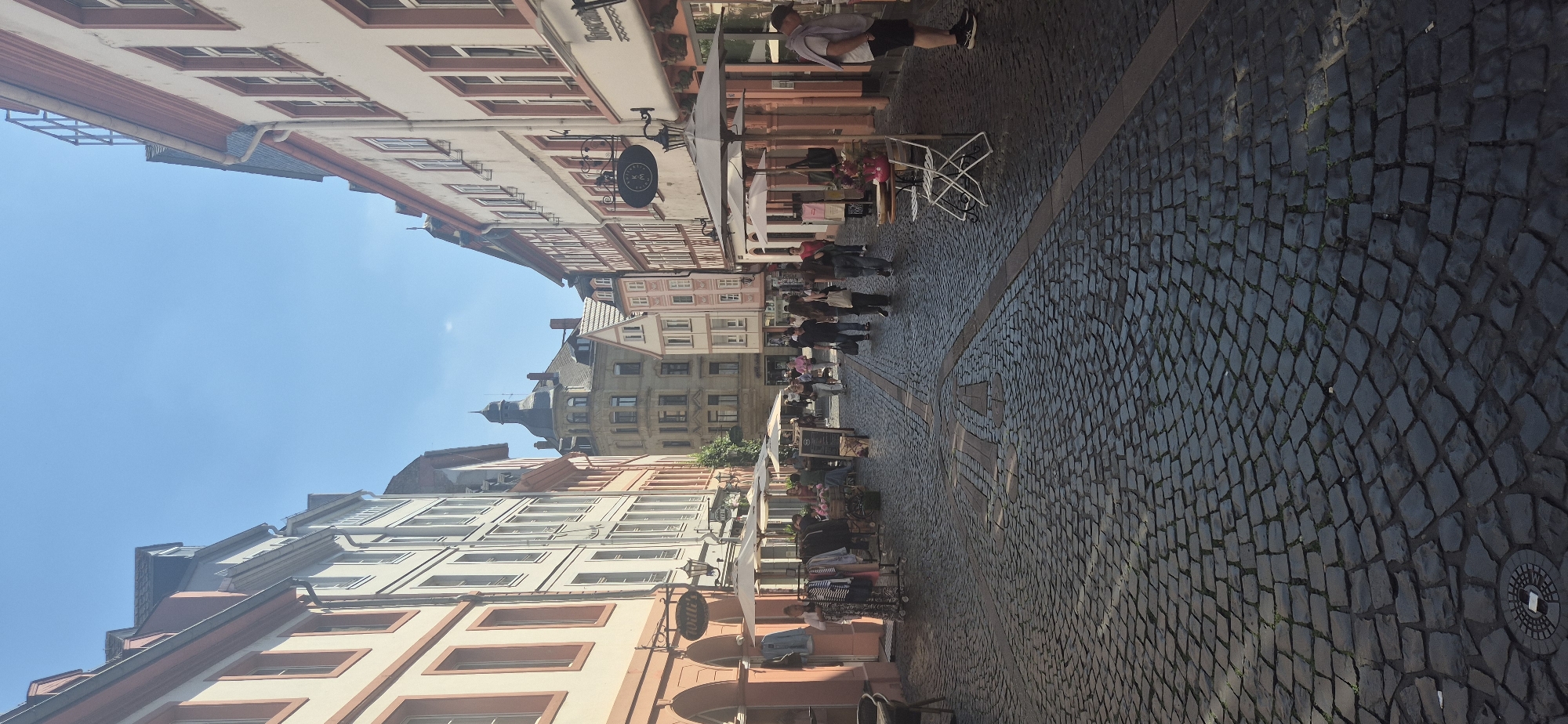 This is a vertical, eye-level, medium shot of the Schustergasse street in Mainz, Germany on a sunny day. The street is paved with cobblestones and lined with narrow buildings featuring traditional European architecture. The buildings have light-colored facades with red trim around the windows and rooflines. Several shops are open, with awnings extended and clothing racks displayed outside. People are walking along the street, some browsing the shops, while others are seated at outdoor tables. The sky is clear and blue. The overall impression is one of a bustling European city street with a marketplace atmosphere.