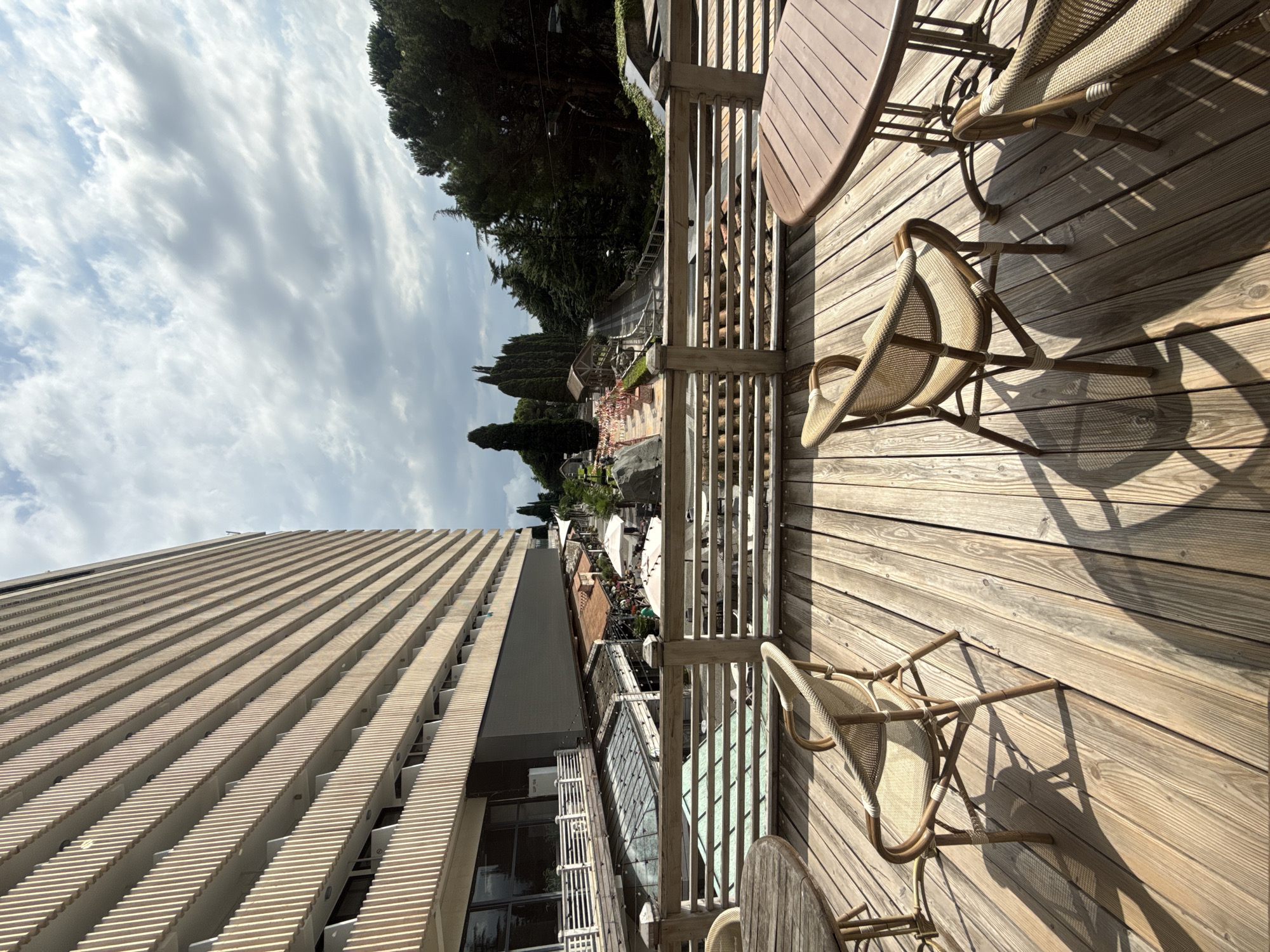 An outdoor seating area with wooden tables and chairs on a wooden deck overlooks a park-like area with trees, a walkway, and a building with repeating balcony structures. The sky is partly cloudy. The location is in Yalta, Ukraine.