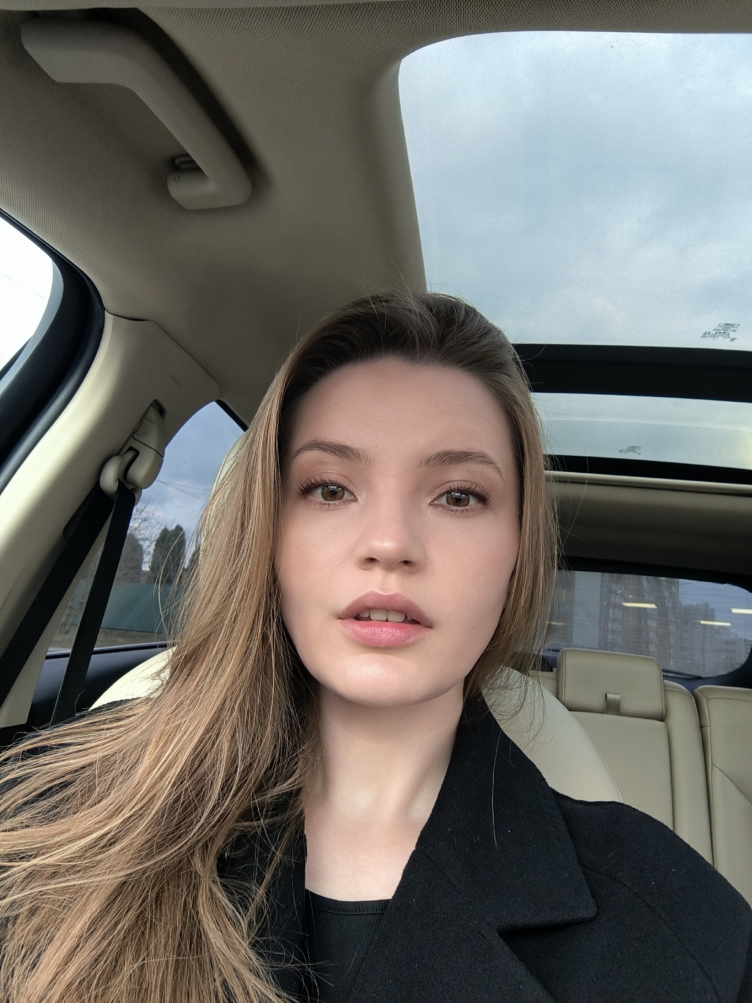 A single female individual is depicted in a self-portrait, seated within the interior of a passenger vehicle. Her face is directed towards the camera with a neutral expression, lips slightly parted. She has long, light brown hair, brown eyes, and fair skin, and is wearing a black collared coat over a dark undergarment.
The vehicle's interior features light-colored upholstery, including the headliner, pillars, and visible rear seats with headrests. A sunroof is visible overhead, showing an overcast sky. A light-colored assist handle is affixed to the ceiling. The left B-pillar shows a black seatbelt mechanism and an "AIRBAG" designation.
The external environment, viewed through the vehicle's windows, suggests an urban or suburban setting in Vinnytsia, Ukraine. Through the left side window, bare trees and a green fence are visible under a light sky. Through the rear window, reflections partially obscure a view of several multi-story buildings, indicative of an urban development.