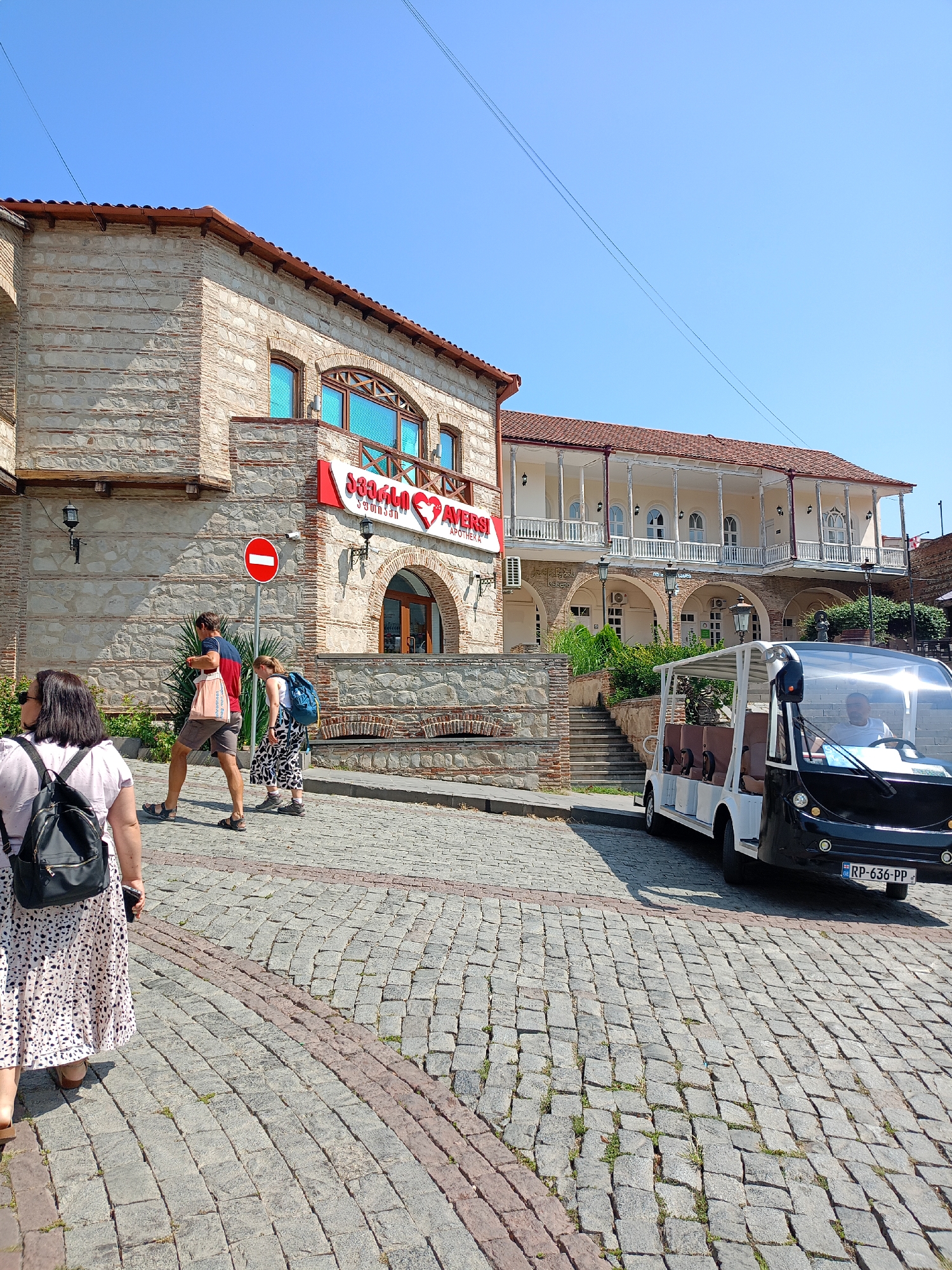 The image depicts a street scene in Sighnaghi, Georgia. A cobblestone street curves through the frame, lined with traditional stone and wooden buildings. On the left, a two-story building with stone walls and a red tiled roof features a sign with Georgian text and a heart symbol. A red and white "no entry" sign is visible on a pole in front of this building.
Several people are walking on the street. In the foreground, a woman with a black backpack and a patterned skirt walks away from the camera. Further back, a man and a woman walk side-by-side, the man carrying a shopping bag.
To the right of the street, a white open-air tour vehicle with a black front is parked. The vehicle has several rows of seats and appears to be a tourist transport. Behind the tour vehicle, another building with a white colonnaded balcony and a red tiled roof is visible. The sky is clear blue. The scene is illuminated by bright sunlight, casting shadows on the ground.