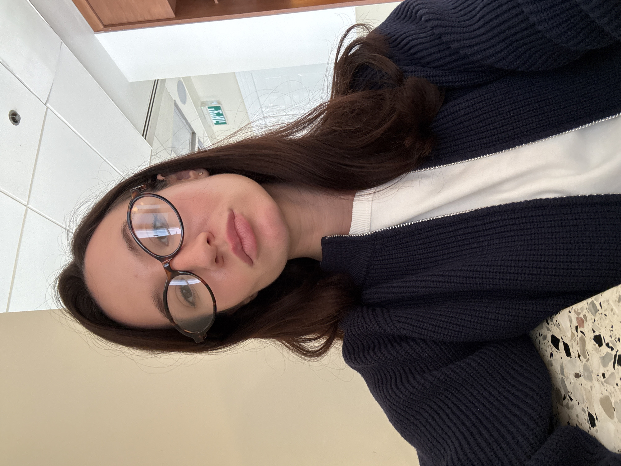 A selfie captures a young woman with long, dark brown hair and fair skin, wearing round, tortoise-shell framed glasses. She looks calmly towards the camera with a neutral expression. Her lips are a natural light pink, slightly parted. She is dressed in a dark blue, ribbed knitted cardigan or jacket, which is unzipped to reveal a white long-sleeved top underneath. The cardigan features a prominent silver zipper. The image is taken from a slightly low angle and is rotated 90 degrees counter-clockwise.
The setting appears to be indoors, likely a public or commercial building in Tel-Aviv, Israel, as indicated by a green sign in the background with white Hebrew text reading "יציאה" (Yetsi'a), which translates to "Exit." The background features a white tiled ceiling with some brown architectural elements above her, and a light-colored wall with horizontal divisions to her side, leading to a brighter hallway or corridor. A speckled, light-colored surface, resembling a terrazzo tabletop, is visible in the lower right foreground. The bright indoor lighting suggests it is daytime. The woman's expression is composed, and the activity is a casual self-portrait.