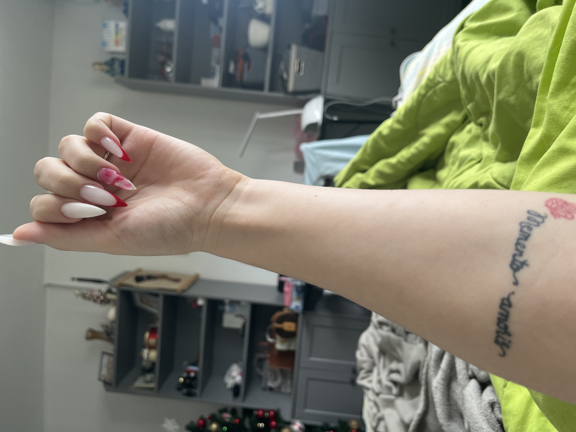 A human arm and hand are positioned horizontally in the foreground. The hand displays long, almond-shaped artificial nails with a red and white color scheme, featuring a marbled design on one nail. The arm has a tattoo near the wrist, displaying the words "Memento amoris" along with a small floral design.
The background is blurred and indistinct, but it appears to show a room interior. Elements visible include shelving with various objects, a green garment, and hints of a Christmas tree.
The image's location is not discernible within Varna, Bulgaria, based on the available visual information.