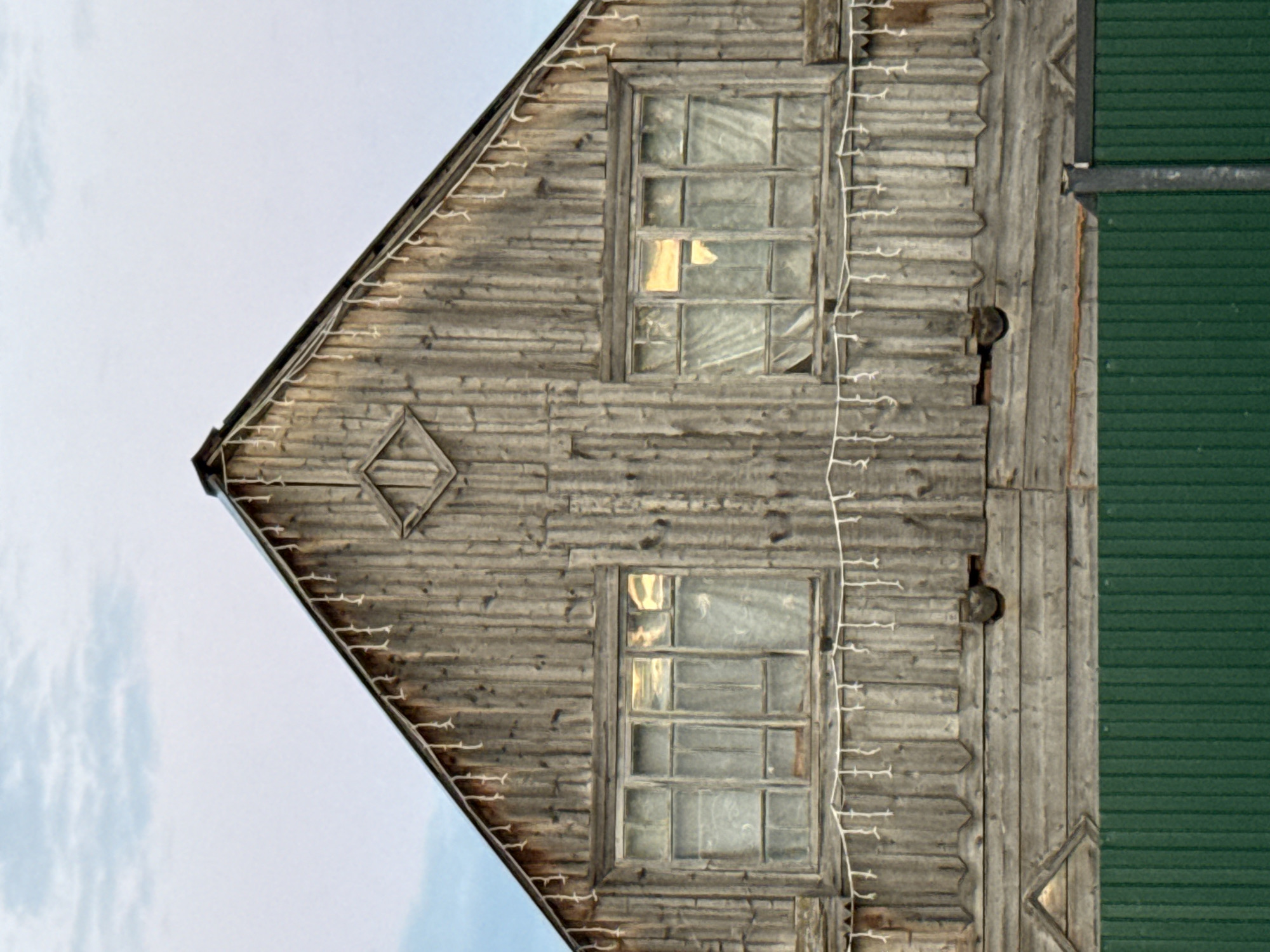 The image shows the facade of an old, wooden house with a gabled roof in the village of Kukui, Russia. The wooden siding is weathered and vertical. Two windows are visible on the upper floor, each with multiple panes. White string lights are draped across the facade below the windows. A diamond-shaped decorative element is centered above the windows, on the gable. Below the main wooden structure is a green metal fence. The sky above is overcast with hints of blue. There are no people or animals visible in the image.