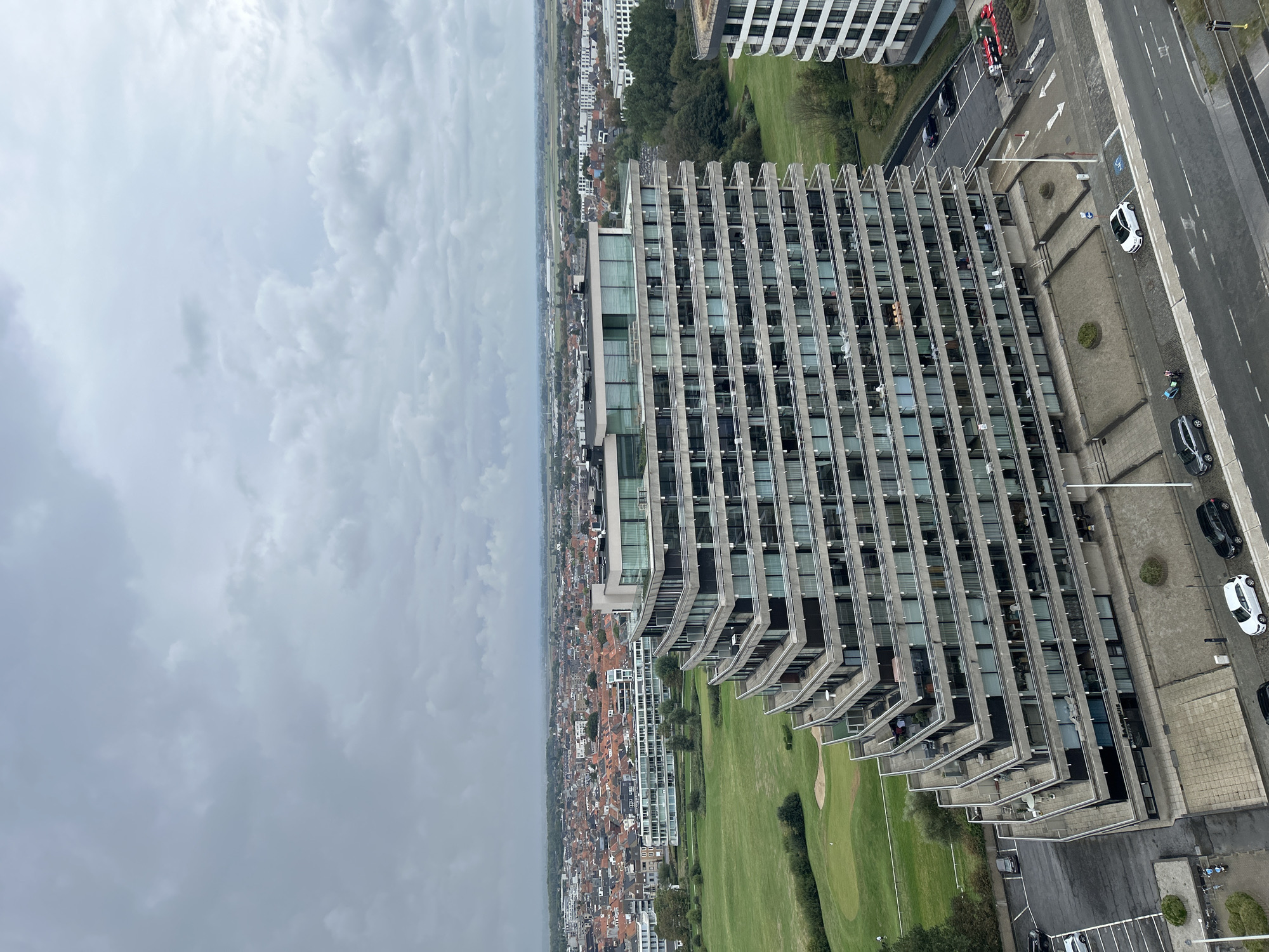 The image is an aerial view of a modern apartment building with a stepped, terraced facade, located in Ostend, Belgium. The building is situated next to a green golf course and overlooks a cityscape with numerous red-tiled roofs and other residential and commercial buildings. A cloudy, overcast sky dominates the upper portion of the frame. The foreground shows a paved area with parked cars and a street with traffic markings. The building's facade is characterized by horizontal bands of balconies and numerous windows. The overall scene depicts an urban coastal environment.