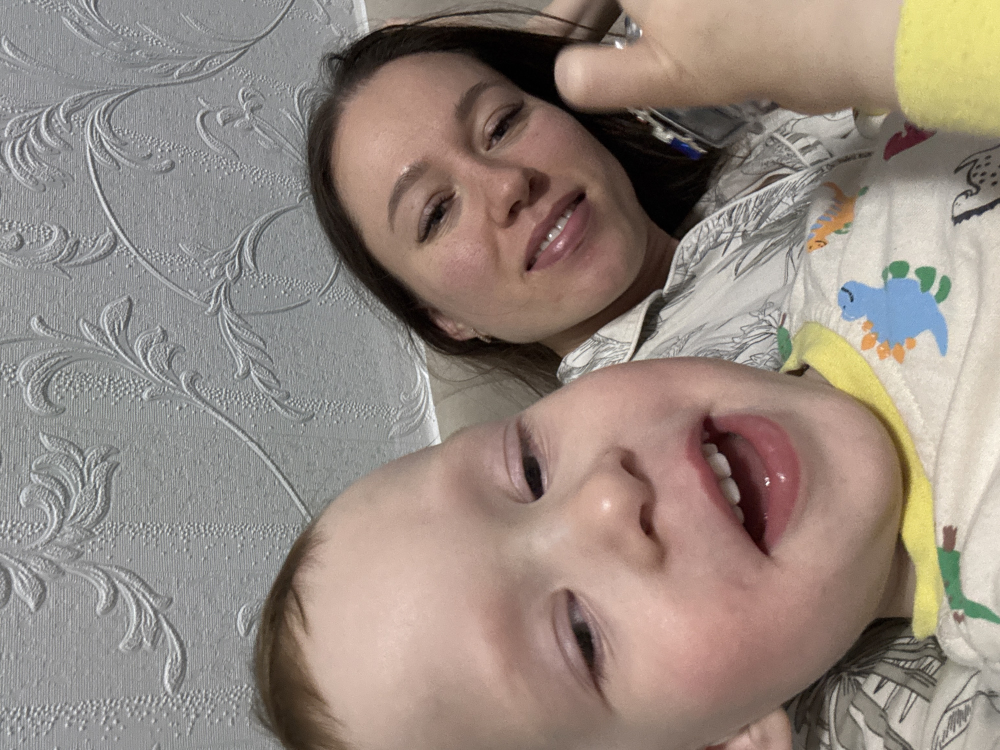 A close-up, upward-angled image captures a woman and a young child lying down together, likely taken as a selfie. The woman, with long dark hair, is smiling gently, resting her head on a surface. She wears a light-colored shirt with a grey leafy pattern, and a ring is visible on her right hand near her head.
Below and in front of the woman is a baby or toddler, also smiling widely with an open mouth, revealing several small teeth. The child has light-colored hair and is dressed in a light-colored top patterned with colorful dinosaurs, such as a blue stegosaurus and an orange brachiosaurus, alongside green foliage. A bright yellow fabric is visible near the child's face.
The background consists of light grey wallpaper featuring an embossed floral or leafy design. The subjects appear content and happy, conveying a warm and affectionate atmosphere in this indoor setting, located in Ferma, Russia. No visible text is present in the image.