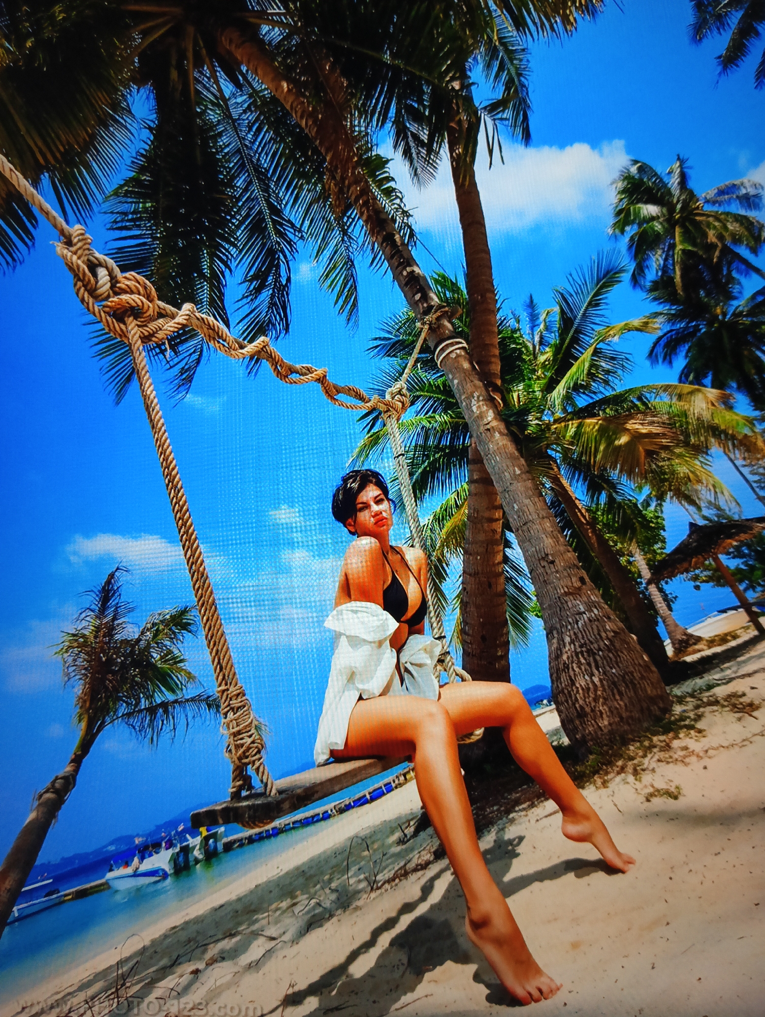 A tanned woman with short dark hair is seated on a wooden swing, under a tropical sky and among palm trees. She is wearing a black bikini top and a loosely tied white shirt. The swing is suspended by thick ropes from the trees. The setting appears to be a beach with sand visible at the bottom of the frame. In the distance, there is water with boats. There is an internet watermark across the bottom of the scene. The scene is typical of the Nha Trang Ward, Vietnam, where beach tourism is present.