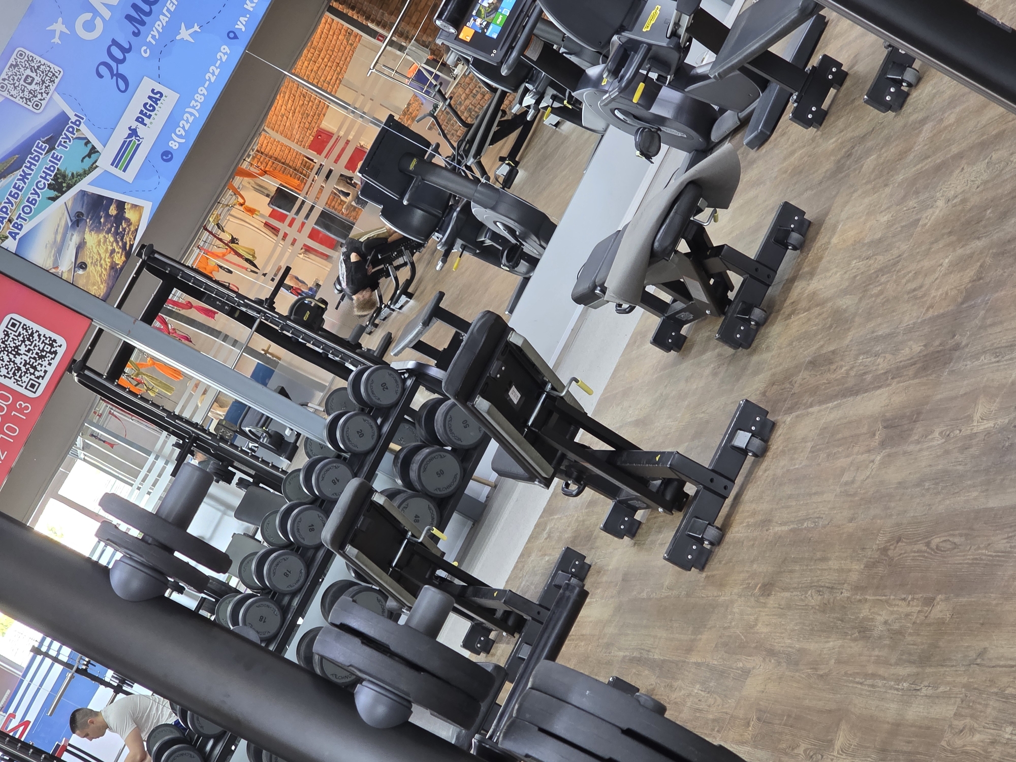 The image shows a gym interior with various exercise equipment. A weight rack with dumbbells is visible in the foreground, along with a leg press machine. Further in the background, there is a seated leg curl machine and other exercise equipment. A man is visible near the weight rack, seemingly occupied with gym activities. Outside the window, there is a view of a building facade. A large advertisement, mentioning "PEGAS TOURISTIK" and "8(922)389-22-29," is attached to the wall, which indicates the gym's location in Russia, specifically, Chaikovsky at the address ул. Ка. The overall impression is that of a well-equipped fitness center.