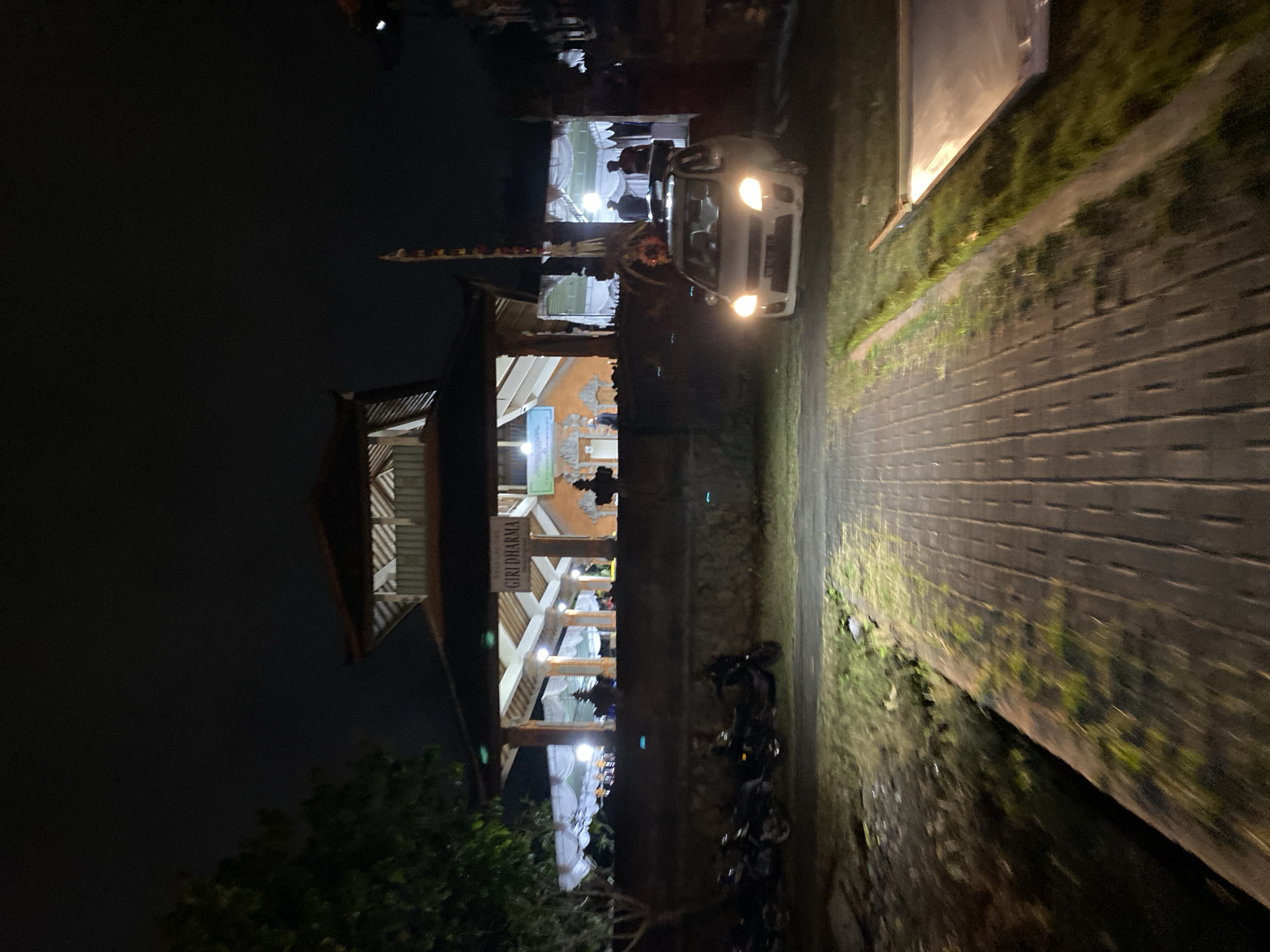 The image depicts an outdoor scene at night, likely in Kuta Selatan, Indonesia. In the background, a building with a prominent roof structure is illuminated, featuring the text "GIRI DHARMA" on a sign. White tents are set up in front of the building, suggesting an event or gathering. Several people are visible under the tents and near the entrance of the building.
In the foreground, a white car with its headlights on is parked on a paved area. To the left of the car, a line of motorcycles is parked along a stone wall. The ground in front of the building is paved with bricks, leading towards the structure. The surrounding area is dimly lit, with some vegetation visible. The overall atmosphere is that of a nighttime event in a community or cultural space.