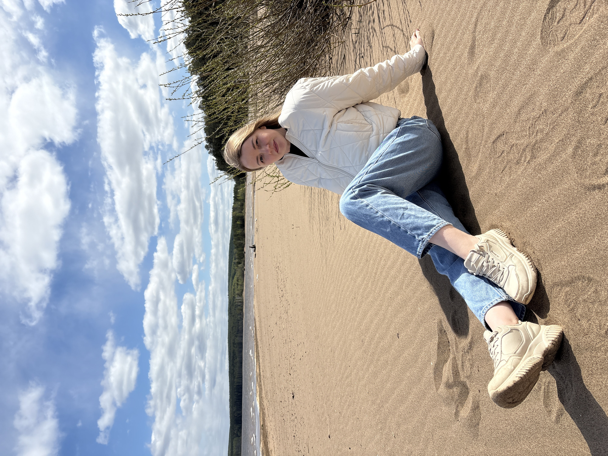 A young woman is lying on a sandy beach in what appears to be Chaikovsky, Russia. She is wearing a white quilted jacket, blue jeans, and beige sneakers. The sky is blue with white clouds. The beach has a line of trees along the shore.
