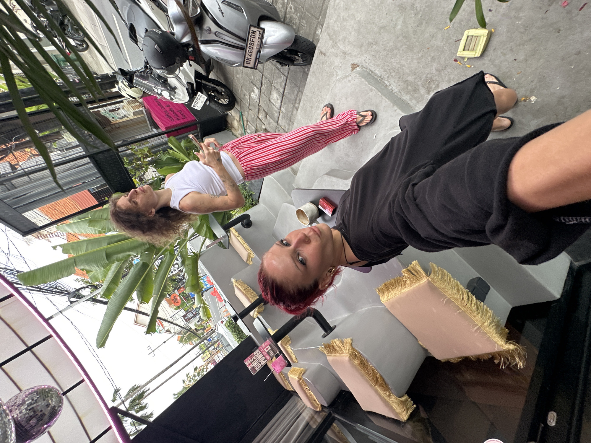 Two women are present in the image, which is set in North Kuta, Indonesia. One woman, in the foreground, has bright red hair and is wearing a black outfit. She is looking towards the camera with a playful expression and is holding her arm out, suggesting she is taking a selfie. She is wearing black flip-flops. The second woman is standing in the background, wearing a white tank top and red and white striped pants. She is holding a smartphone in her hands and looking to her right, away from the camera. She is also wearing flip-flops.
The environment appears to be an outdoor seating area with tiered seating. The seats are covered in a light pink fabric with gold fringe. There are potted plants and palm trees visible, suggesting a tropical location. In the background, there are motorcycles parked along a street. A disco ball hangs from the ceiling structure above. A small table in the middle ground holds a cup and a small box. The overall scene suggests a casual, possibly cafe or bar setting.