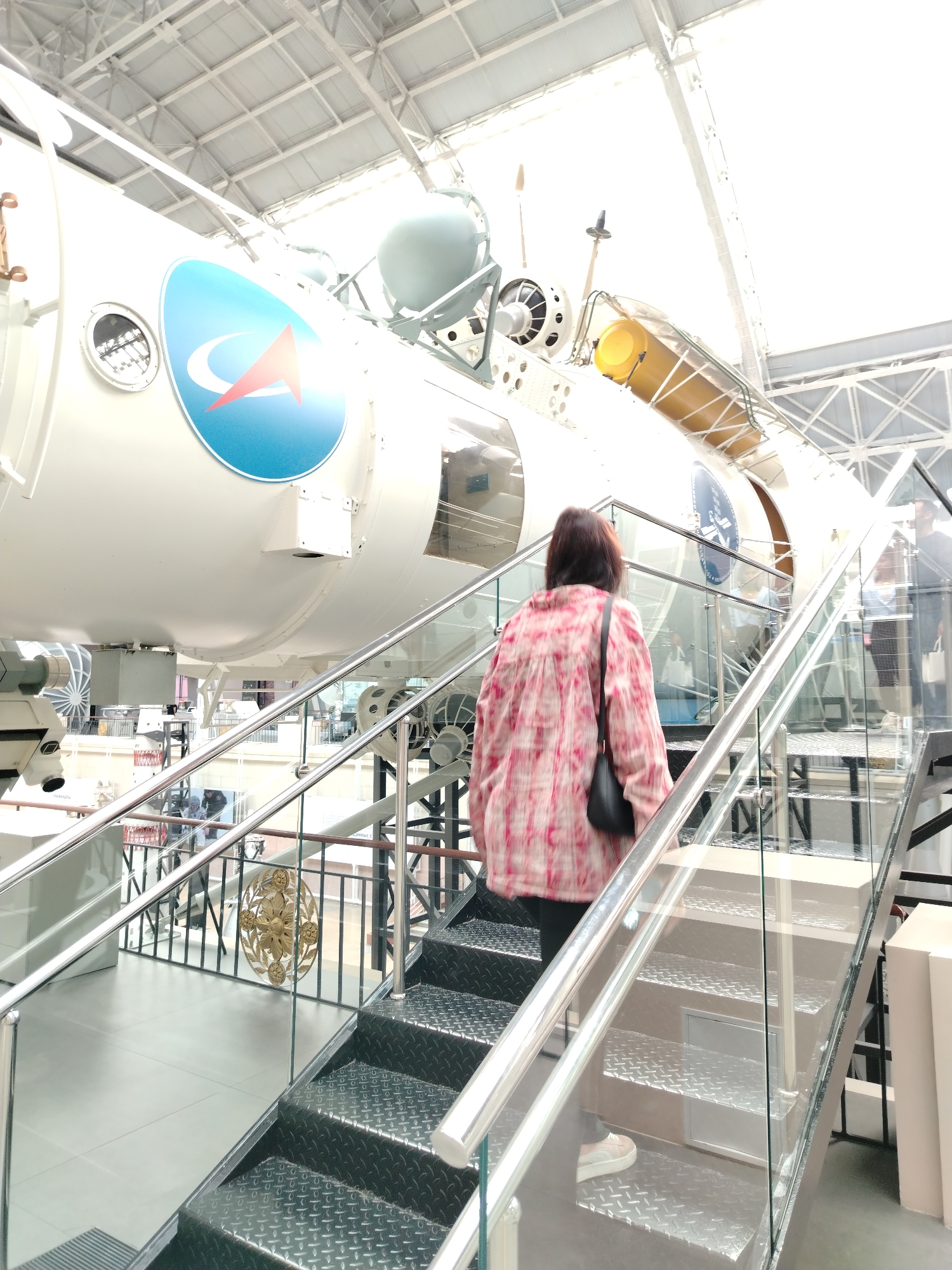 The image shows a woman with dark hair, wearing a pink and white patterned shirt and white sneakers, walking up a metal staircase with glass railings. She is carrying a black purse over her shoulder. The staircase leads to a large white spacecraft on display indoors. The spacecraft has a prominent blue and red logo on its side and various cylindrical components. The environment appears to be a museum or exhibition hall with a high ceiling and industrial-style architecture. There are other people visible in the background, also looking at the spacecraft. This scene is located at the Memorial Museum of Cosmonautics in Moscow, Russia, as evidenced by the presence of the spacecraft and the overall museum setting.